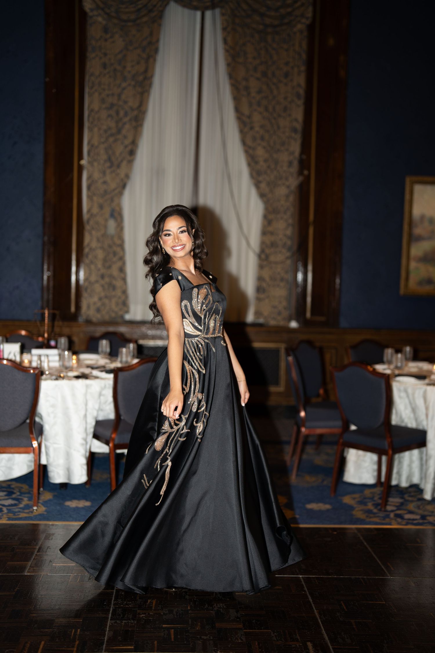 Woman in stunning black and gold embroidered ball gown twirling in elegant blue ballroom venue.