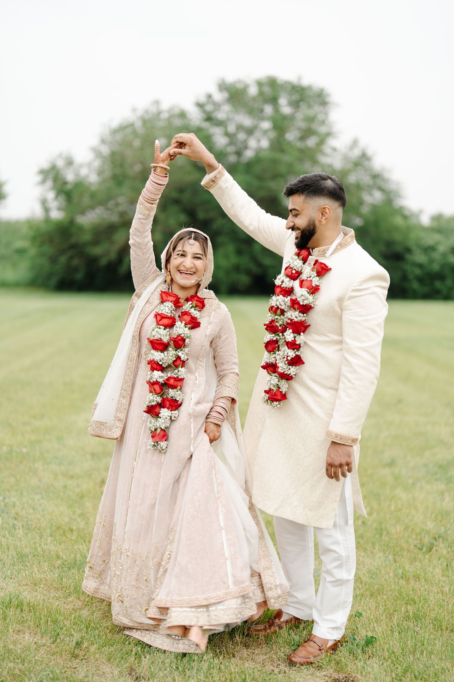 Indian bride and groom in traditional attire with red floral garlands dancing outdoors on green grass.