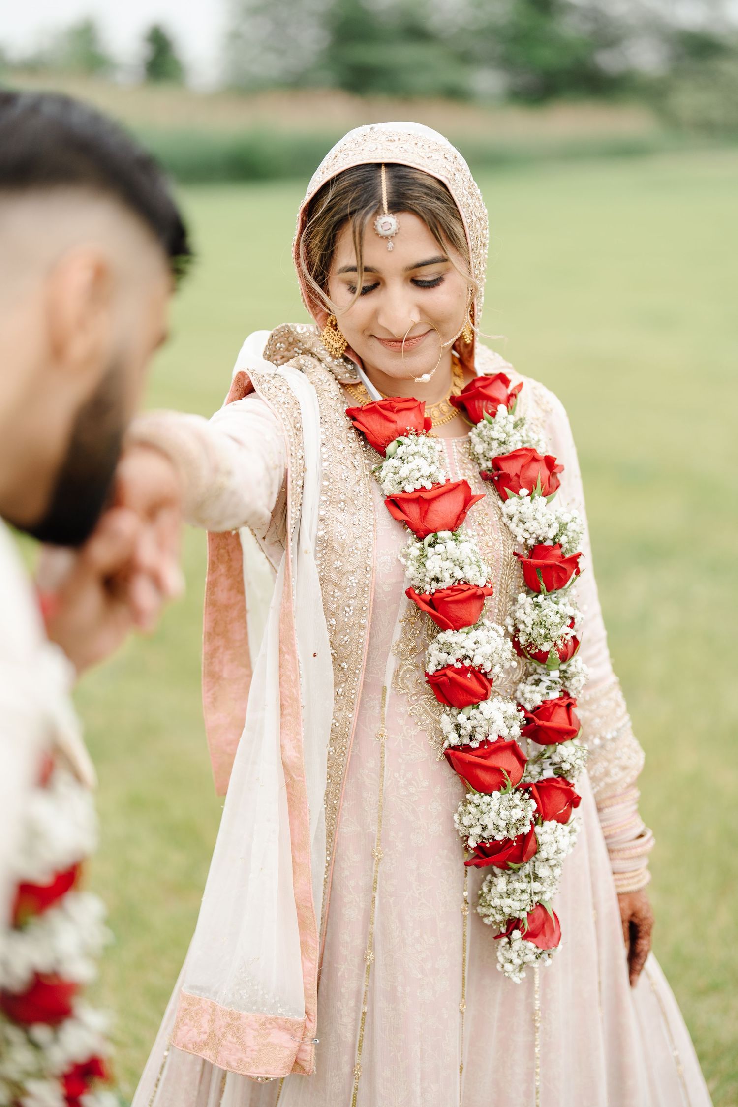 Indian bride in blush lehenga with red rose garland smiling while groom holds her hand outdoors.