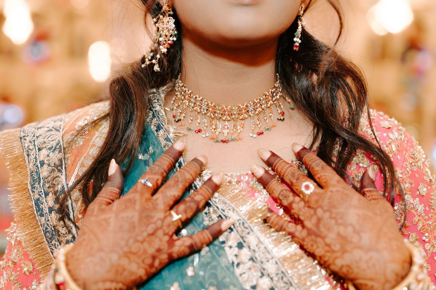 Close-up of Indian bride with mehndi hands, kundan necklace, and earrings in colorful bridal attire.