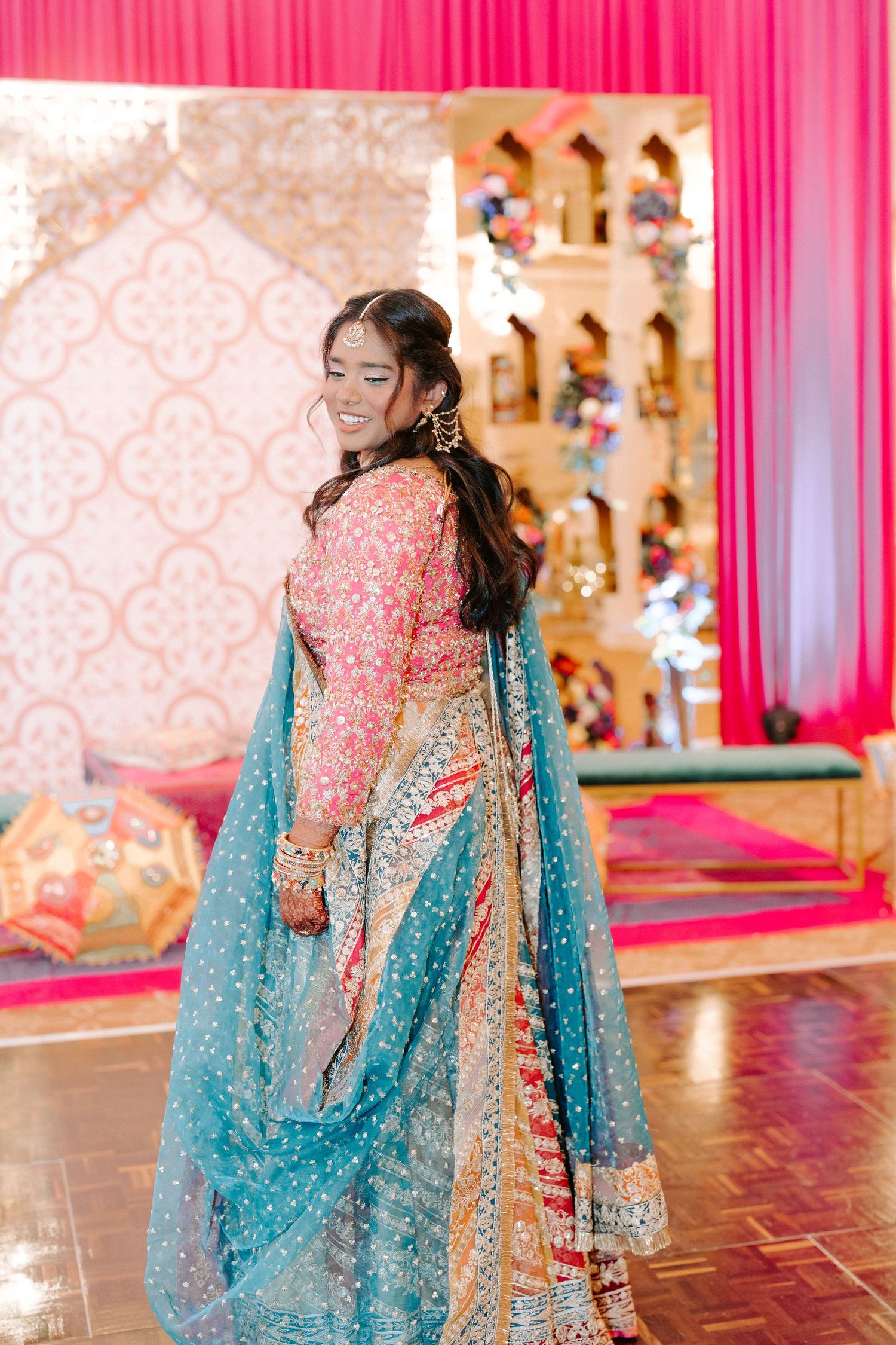 Bride in pink and gold lehenga with teal dupatta smiling at vibrant South Asian wedding.
