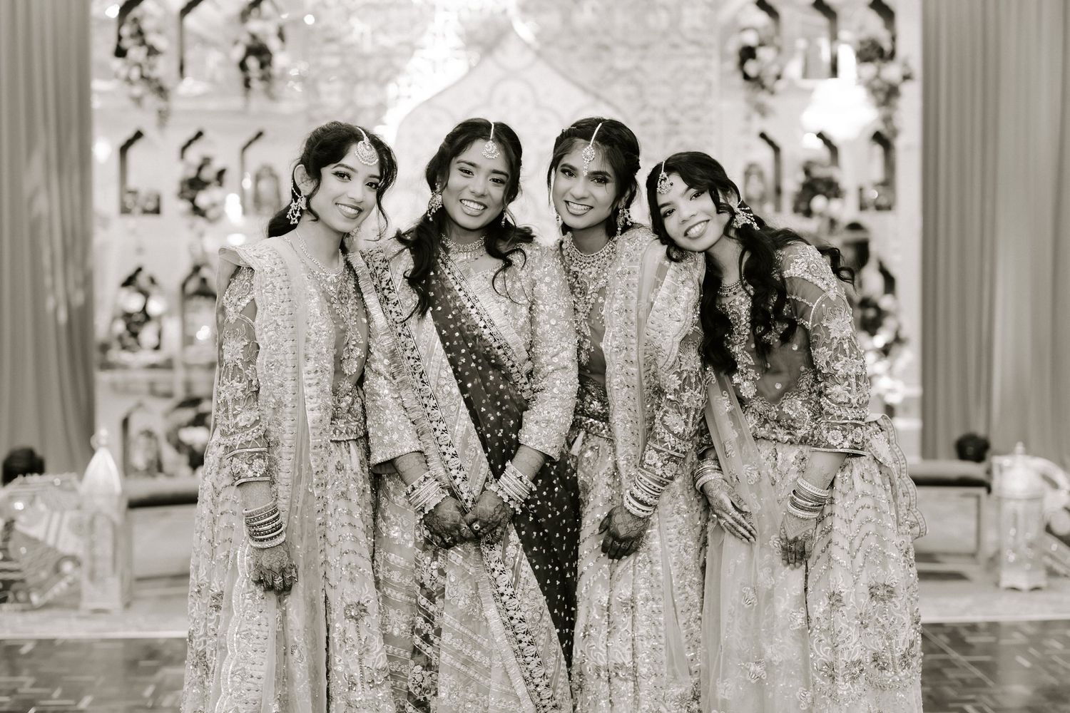 Four women in elegant Indian wedding attire posing together in black and white photo at ceremony.