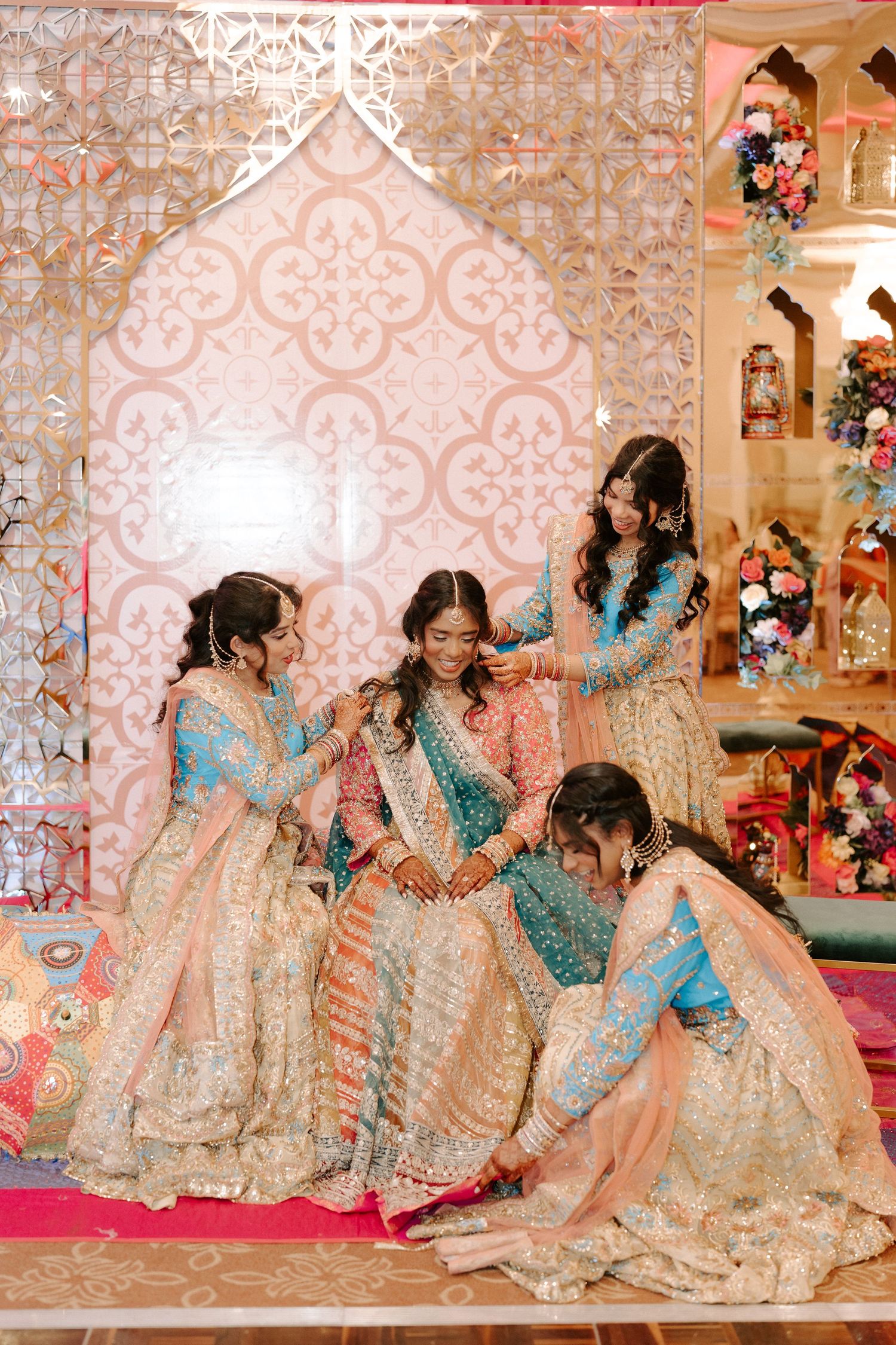 Bridesmaids in blue and gold lehengas help bride prepare at elegant Indian wedding ceremony backdrop.