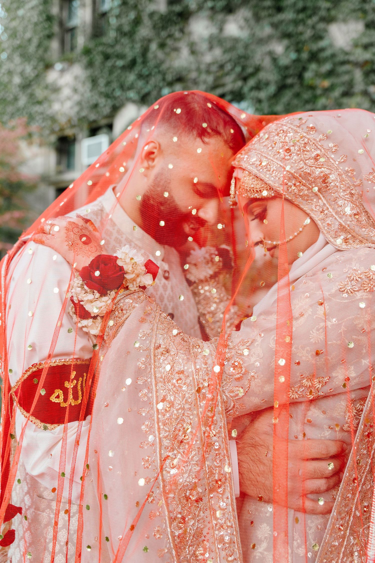 South Asian bride and groom sharing intimate moment under sheer red dupatta veil with Allah calligraphy.