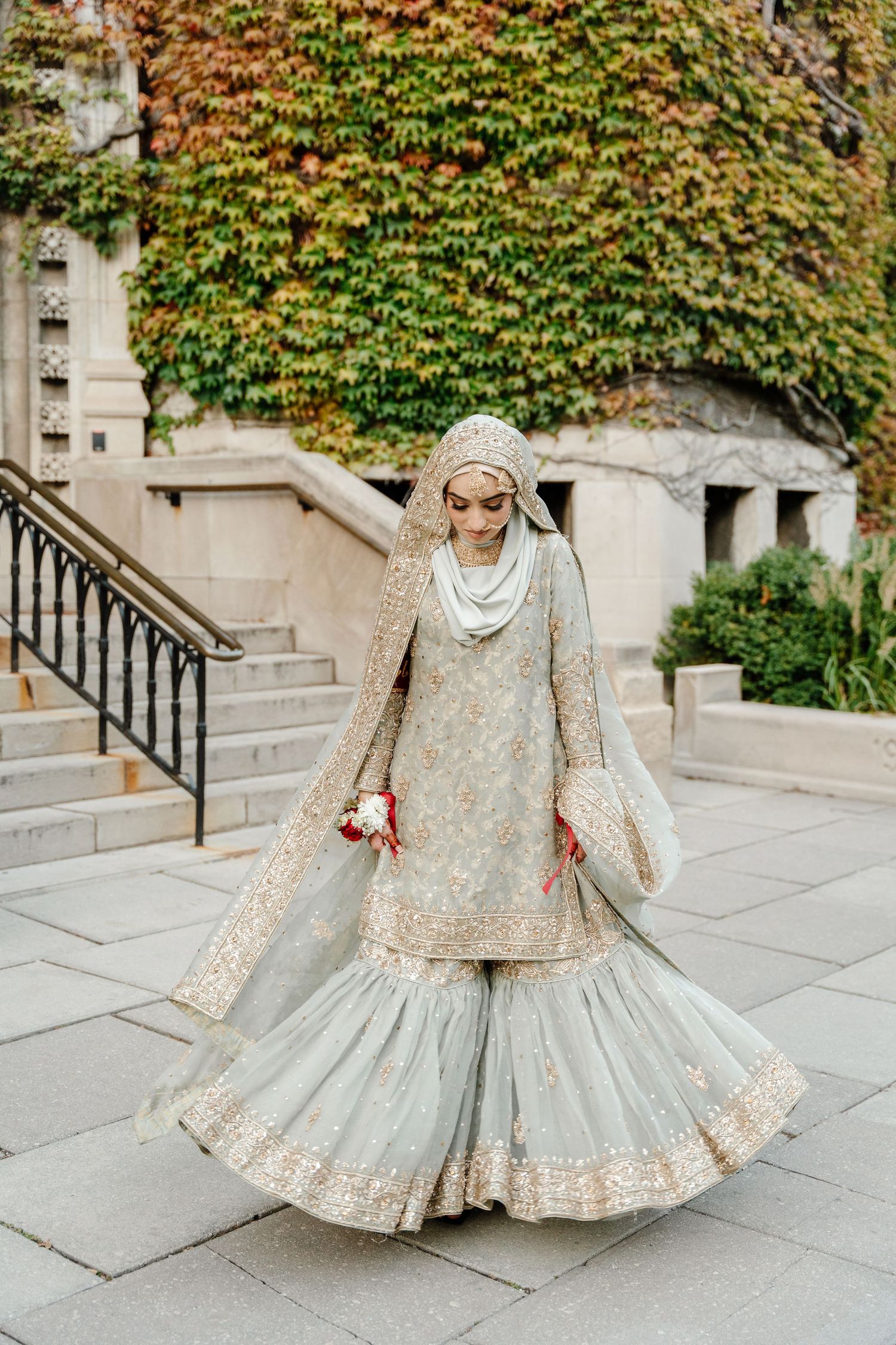 Muslim bride in mint green embroidered sharara suit with hijab twirling outdoors near ivy-covered stone building.