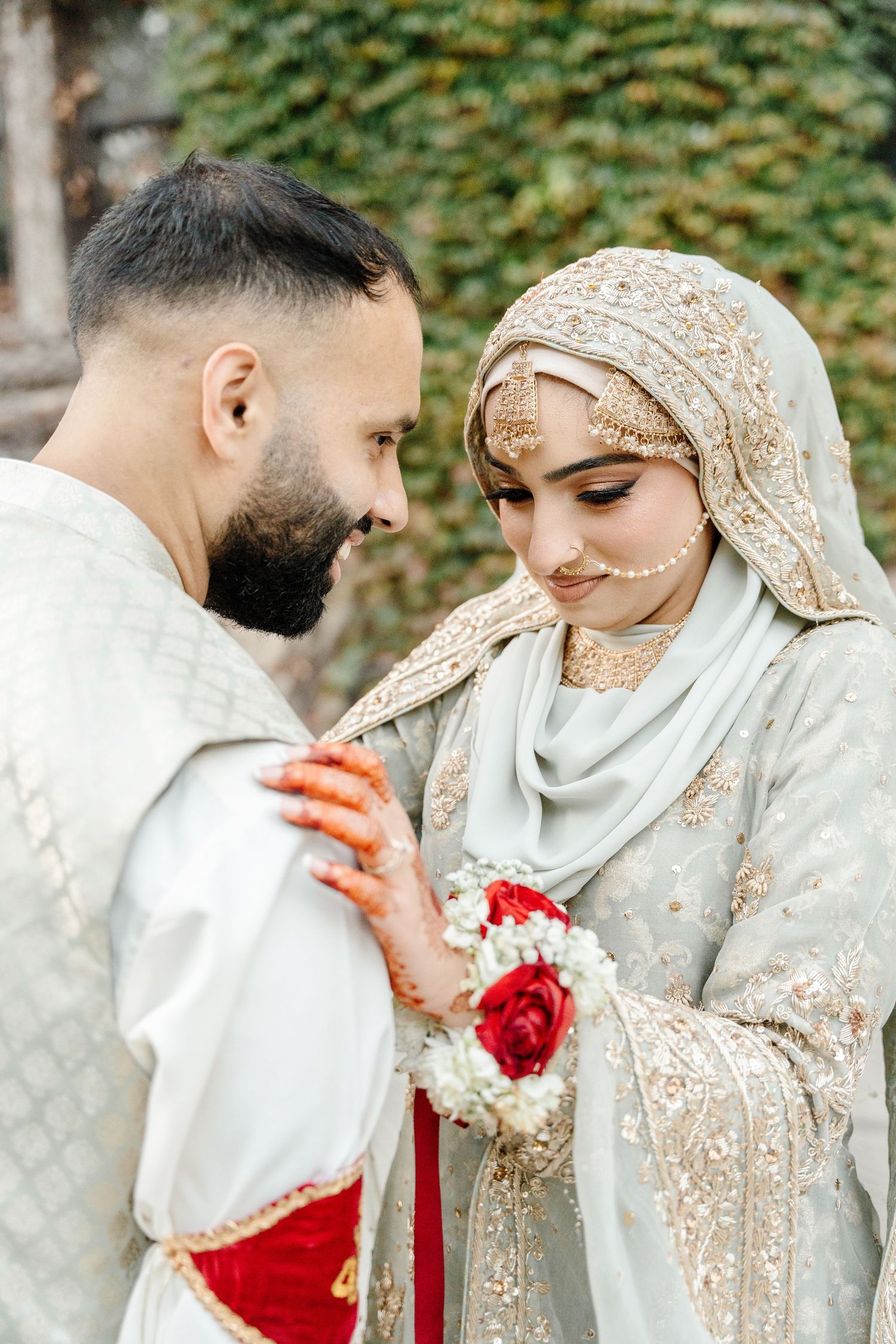 Pakistani bride and groom in traditional embroidered wedding attire sharing tender moment with red rose bouquet.