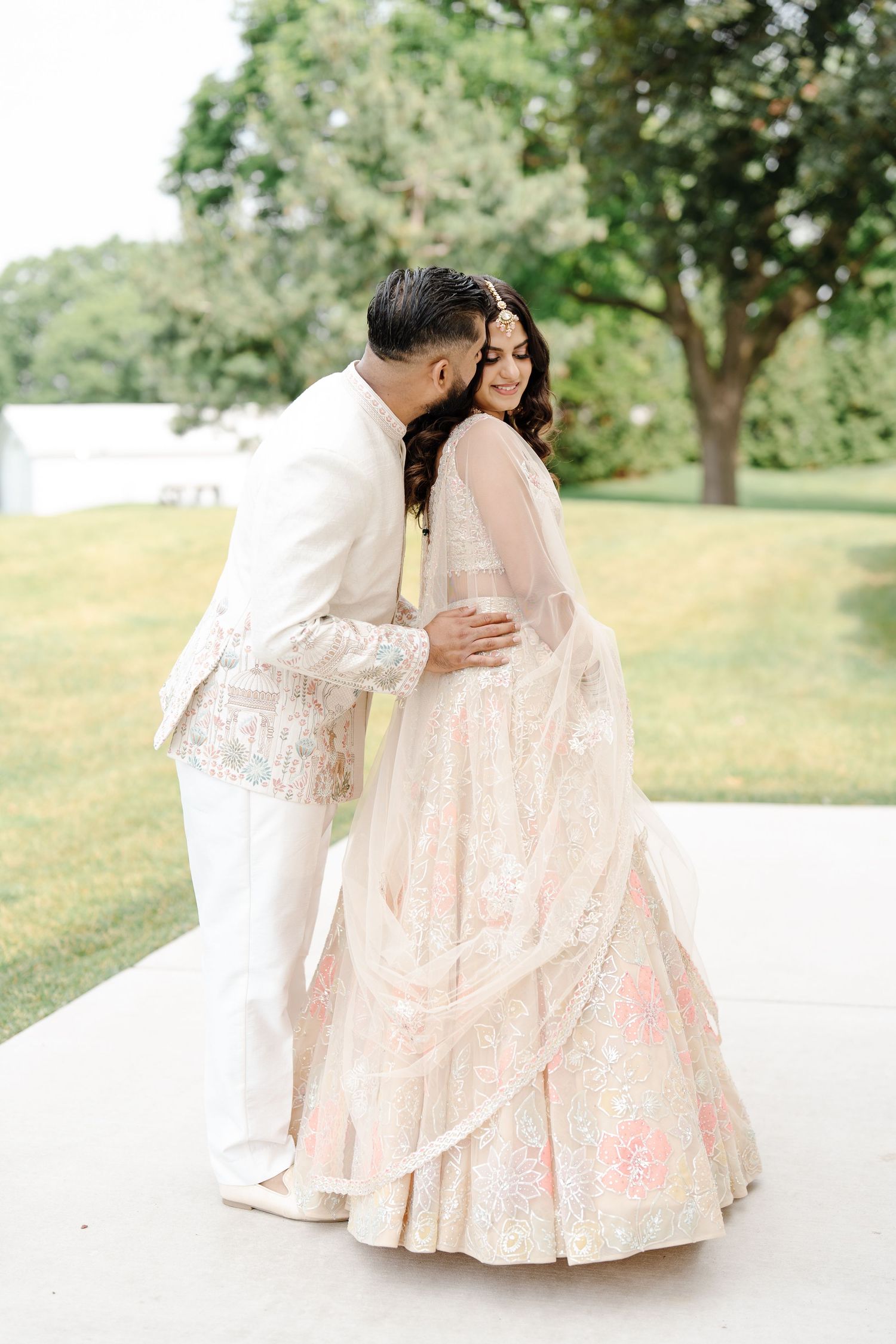 Groom in white sherwani embracing bride in pastel floral lehenga during romantic outdoor wedding portrait.