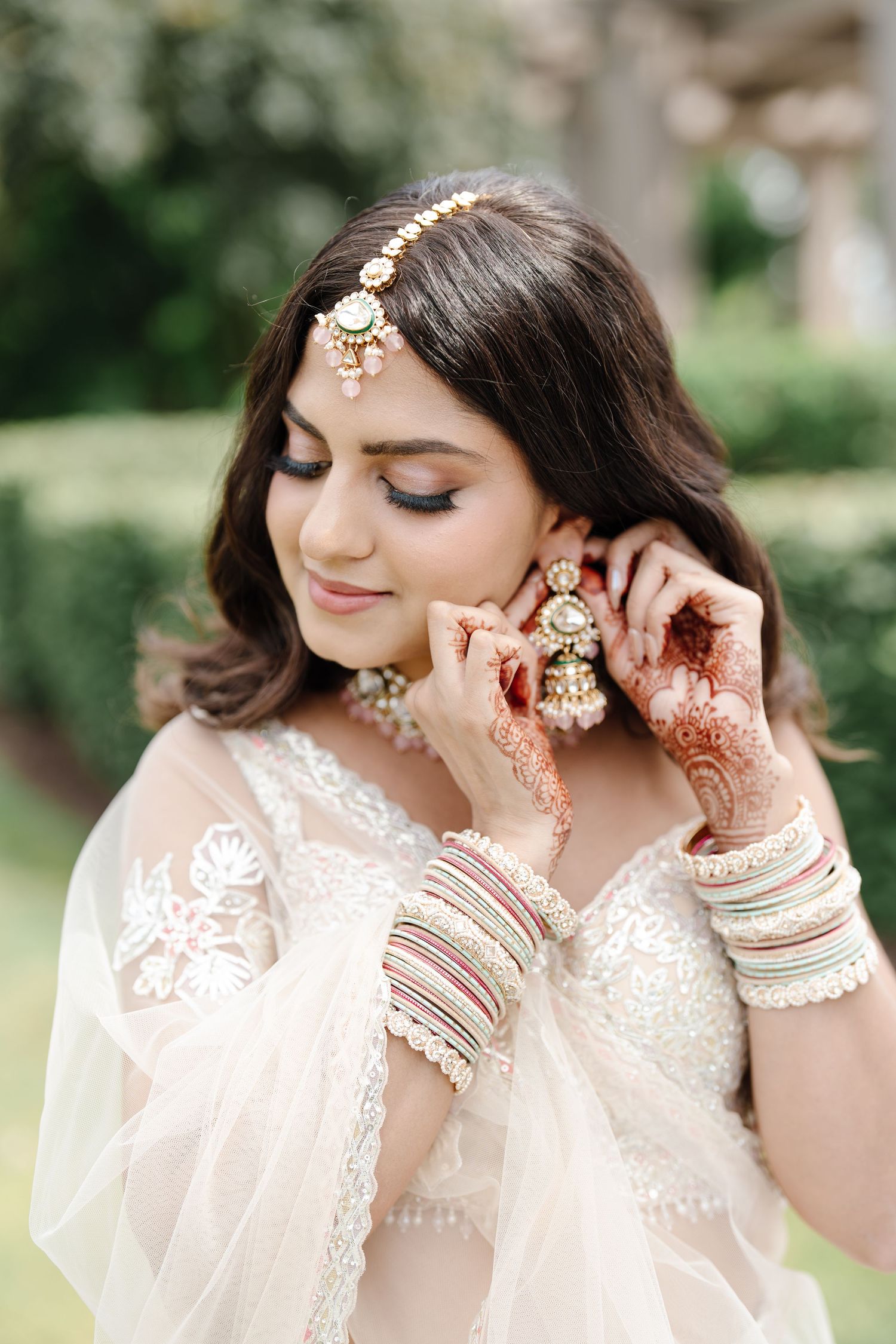 Indian bride adjusting jhumka earrings with mehndi hands, wearing kundan jewelry and white lehenga.