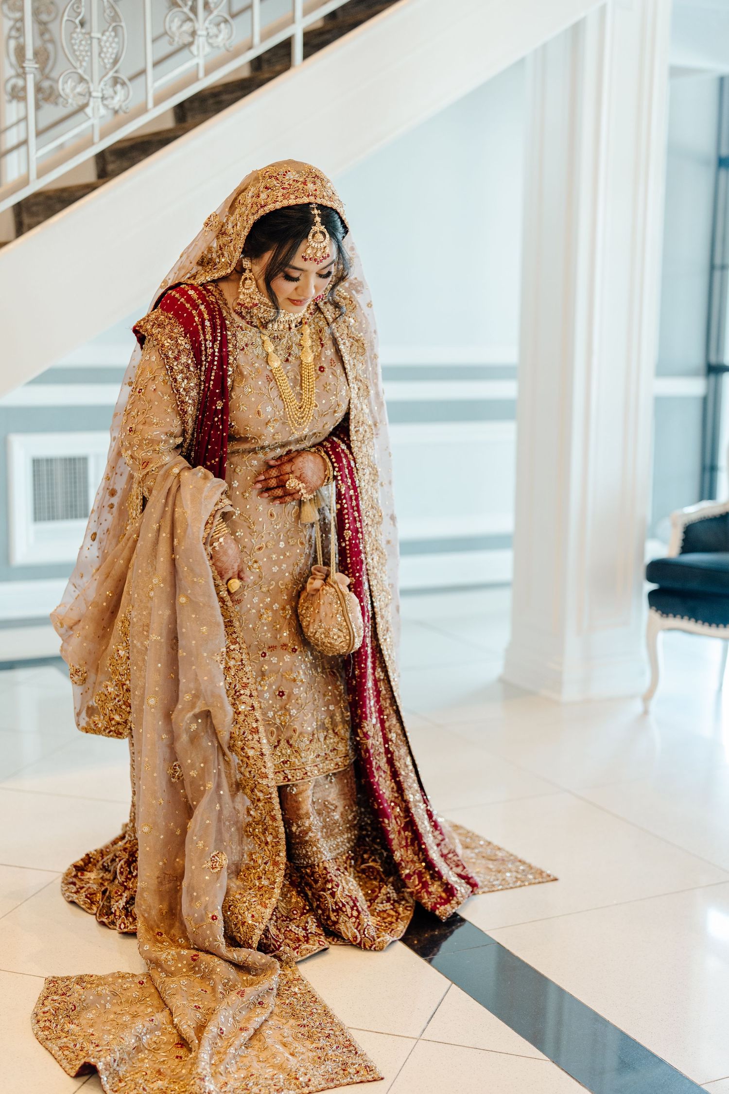 South Asian bride in ornate gold and maroon bridal lehenga with heavy embroidery standing on marble staircase.