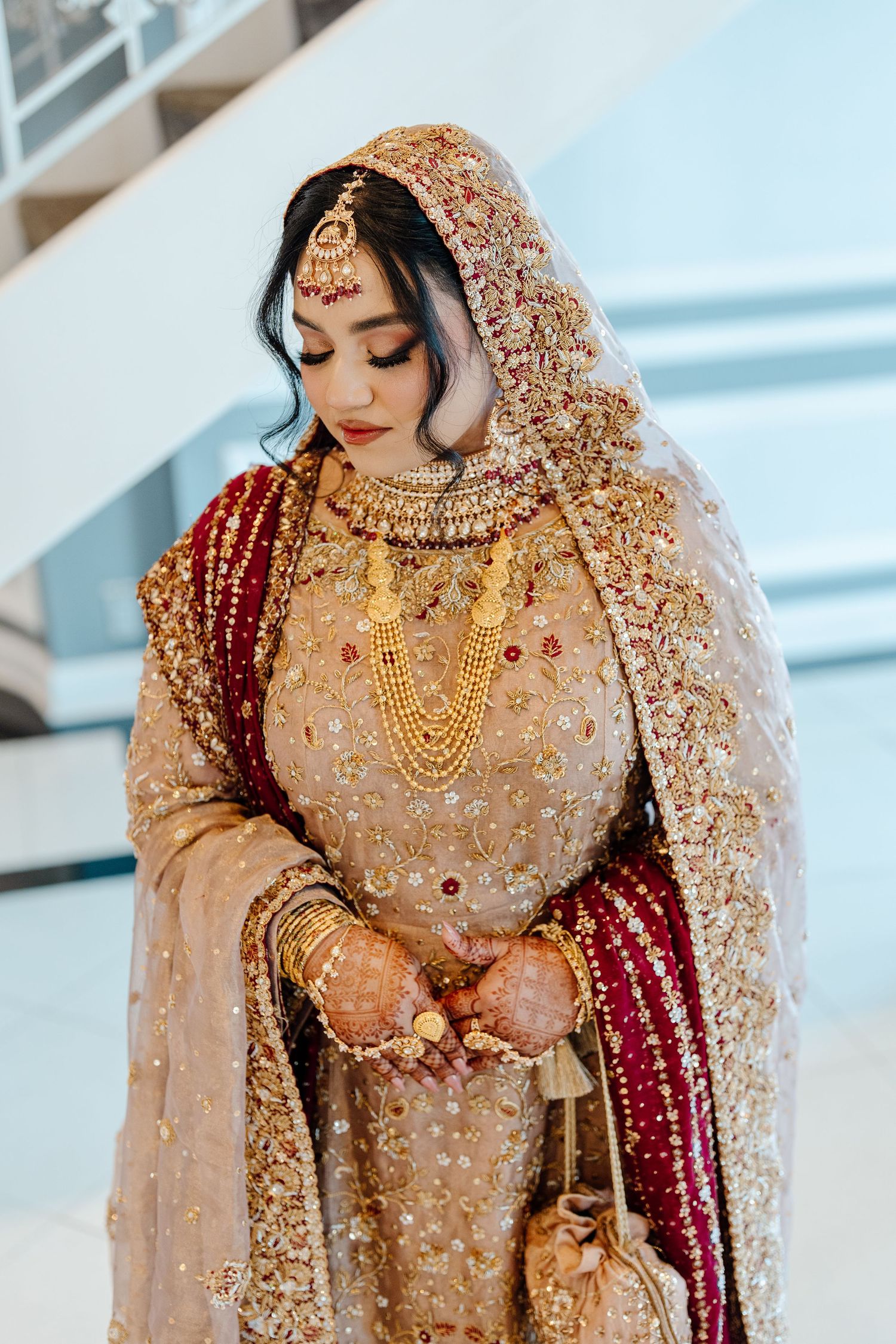 Pakistani bride wearing heavily embroidered gold bridal lehenga with layered gold jewelry and maang tikka indoors.