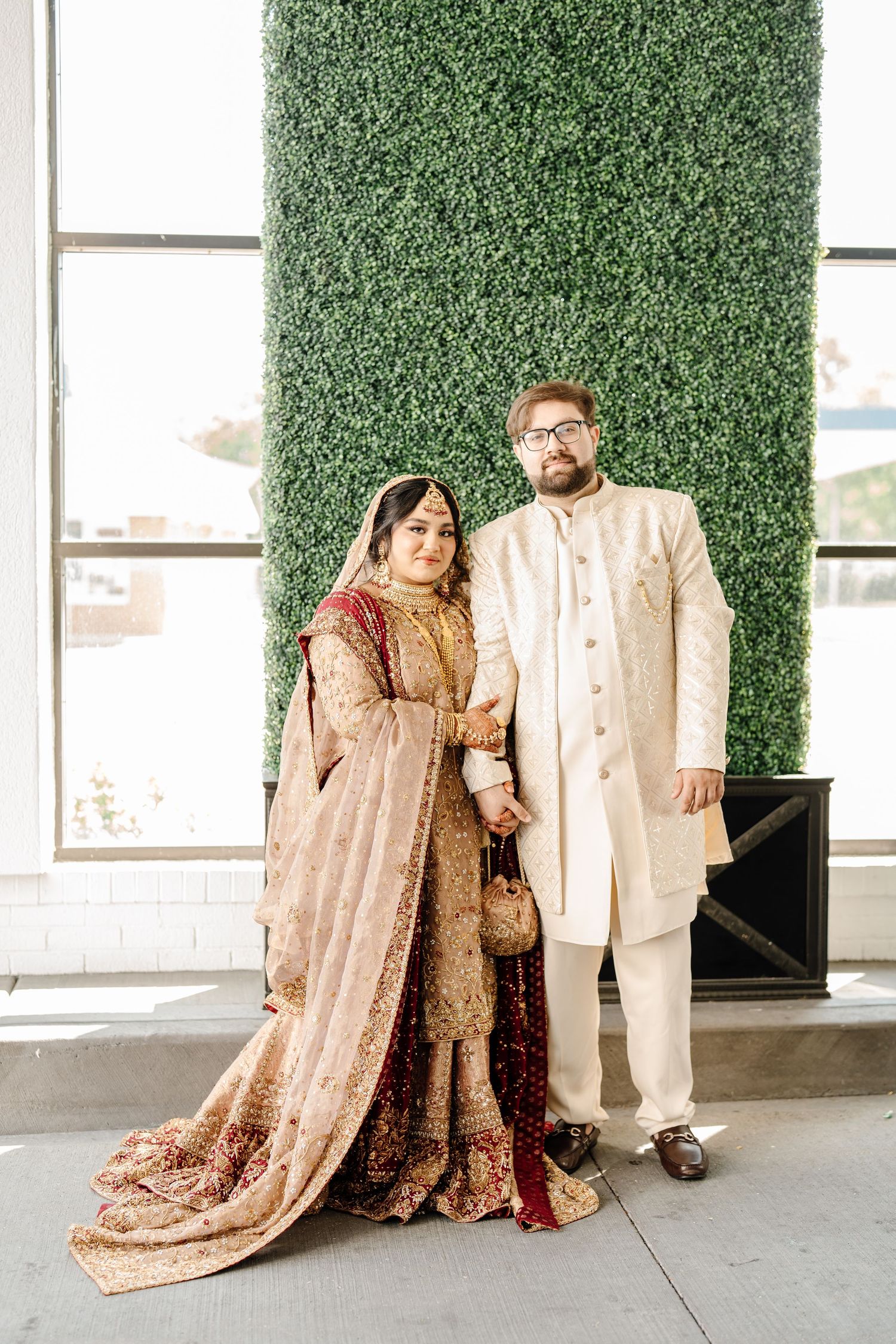 Pakistani bride and groom in traditional gold and cream wedding attire posing before green hedge wall backdrop.
