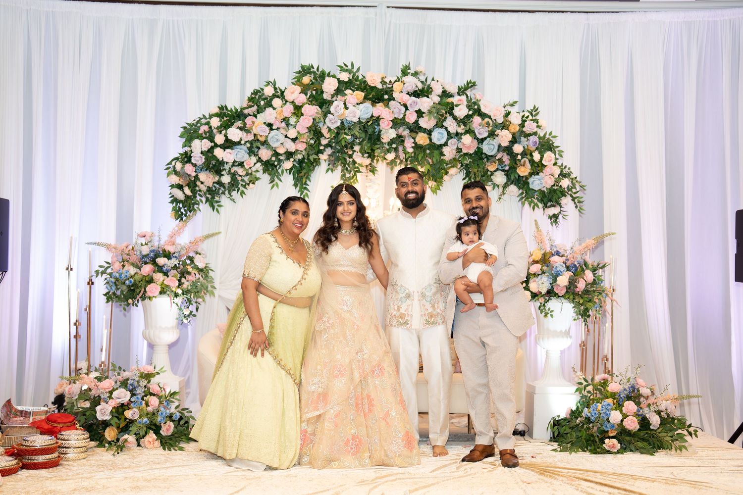 Indian wedding couple with family posing under floral arch with pink and white blooms at reception.