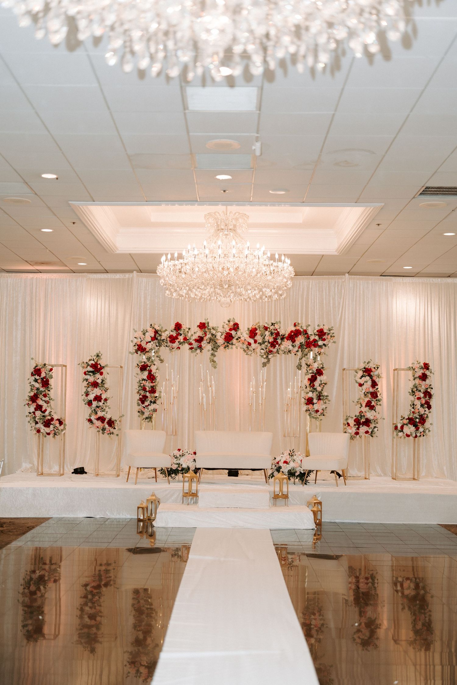 Grand wedding ceremony backdrop with red and white floral arrangements under crystal chandelier.
