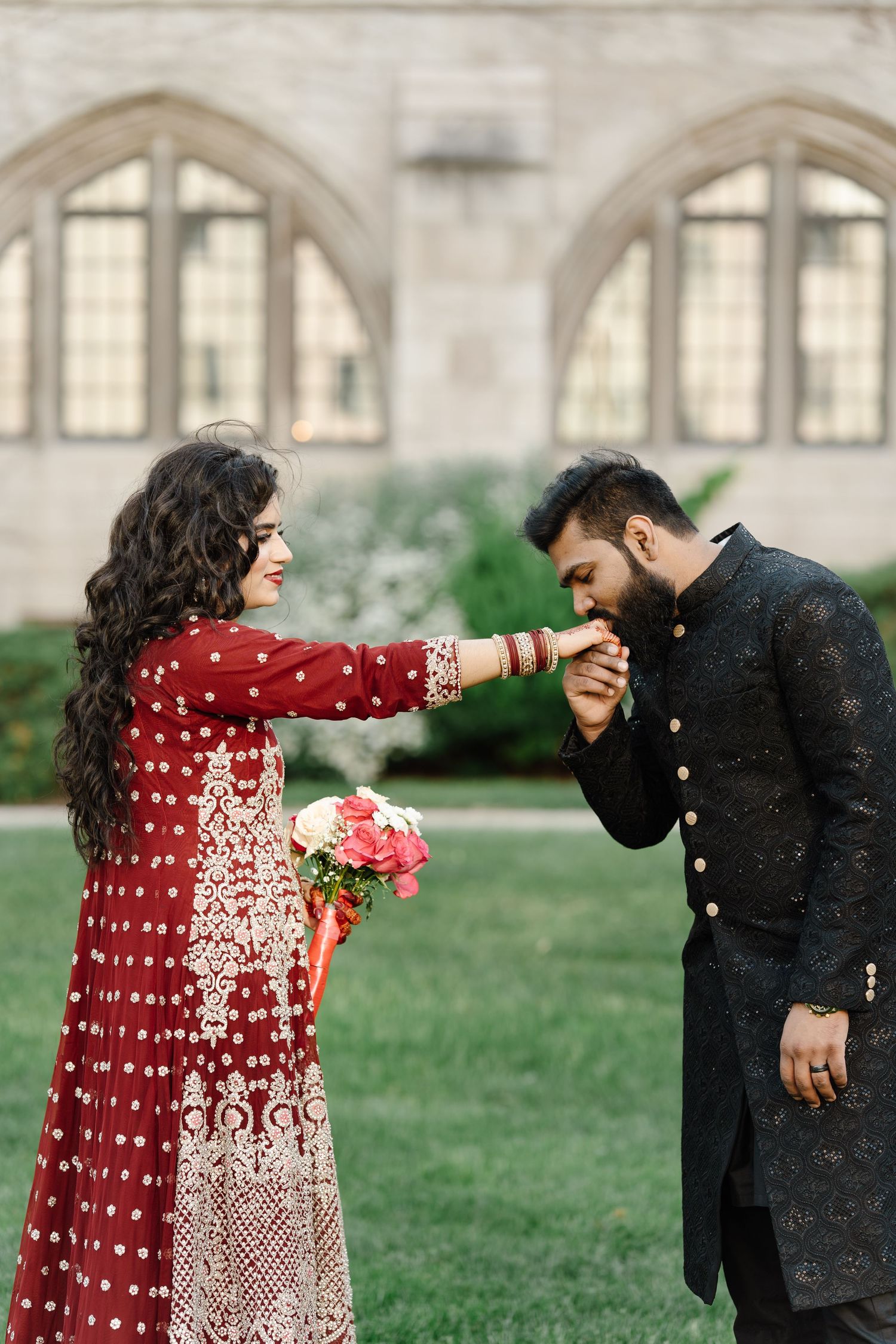 Man in black sherwani kissing woman's hand during South Asian pre-wedding photoshoot at stone building.