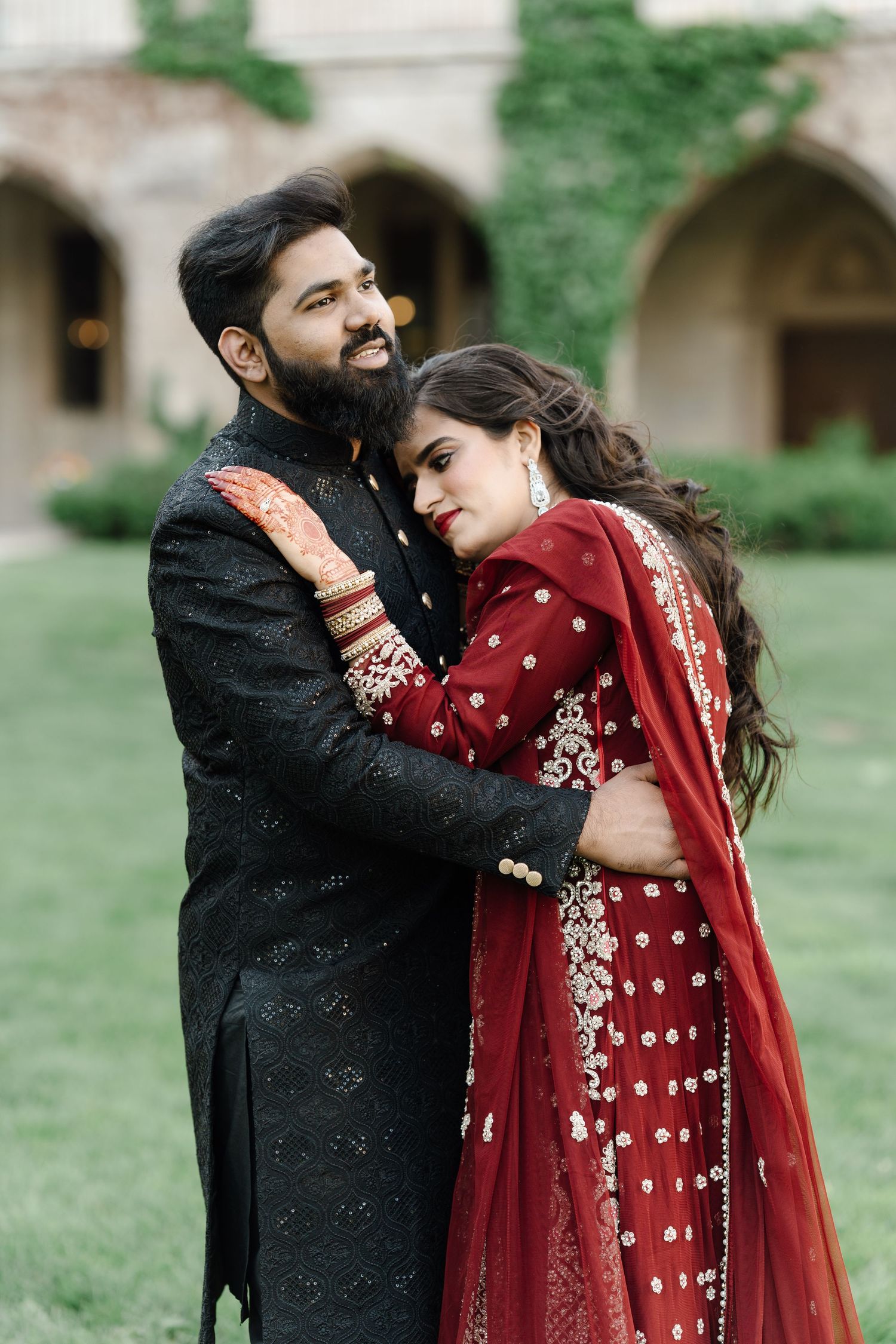 South Asian couple embracing in red and black traditional wedding attire at historic stone courtyard.
