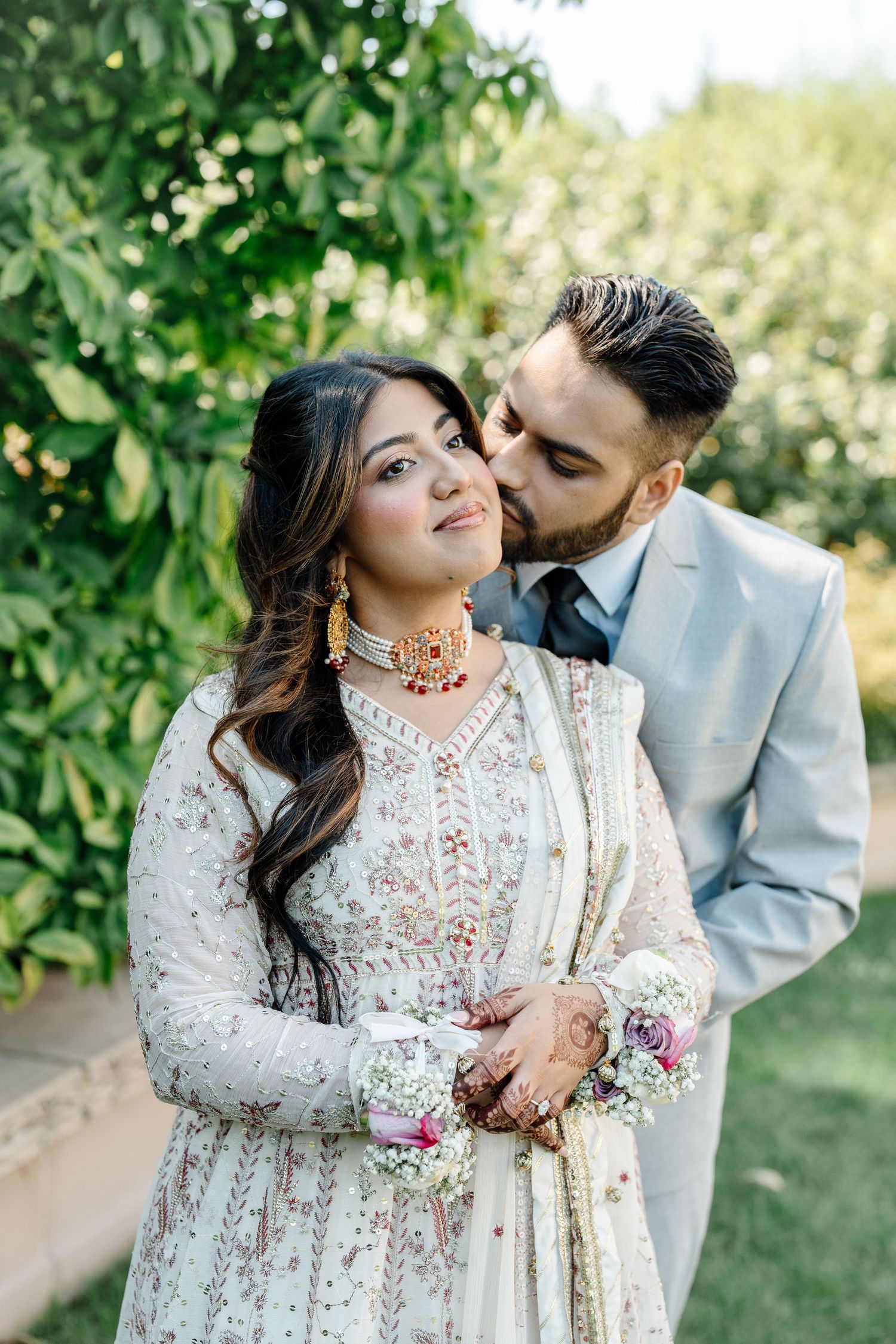 South Asian groom in grey suit kissing bride wearing white embroidered anarkali with ruby choker necklace outdoors.