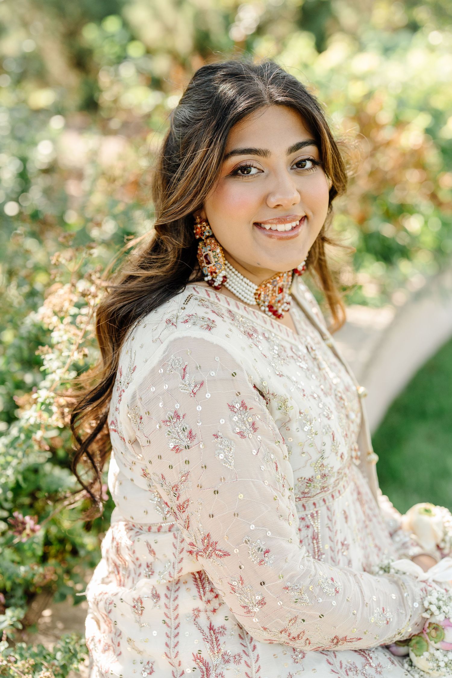 Smiling South Asian bride in white embroidered lehenga with ornate earrings posing outdoors among greenery.