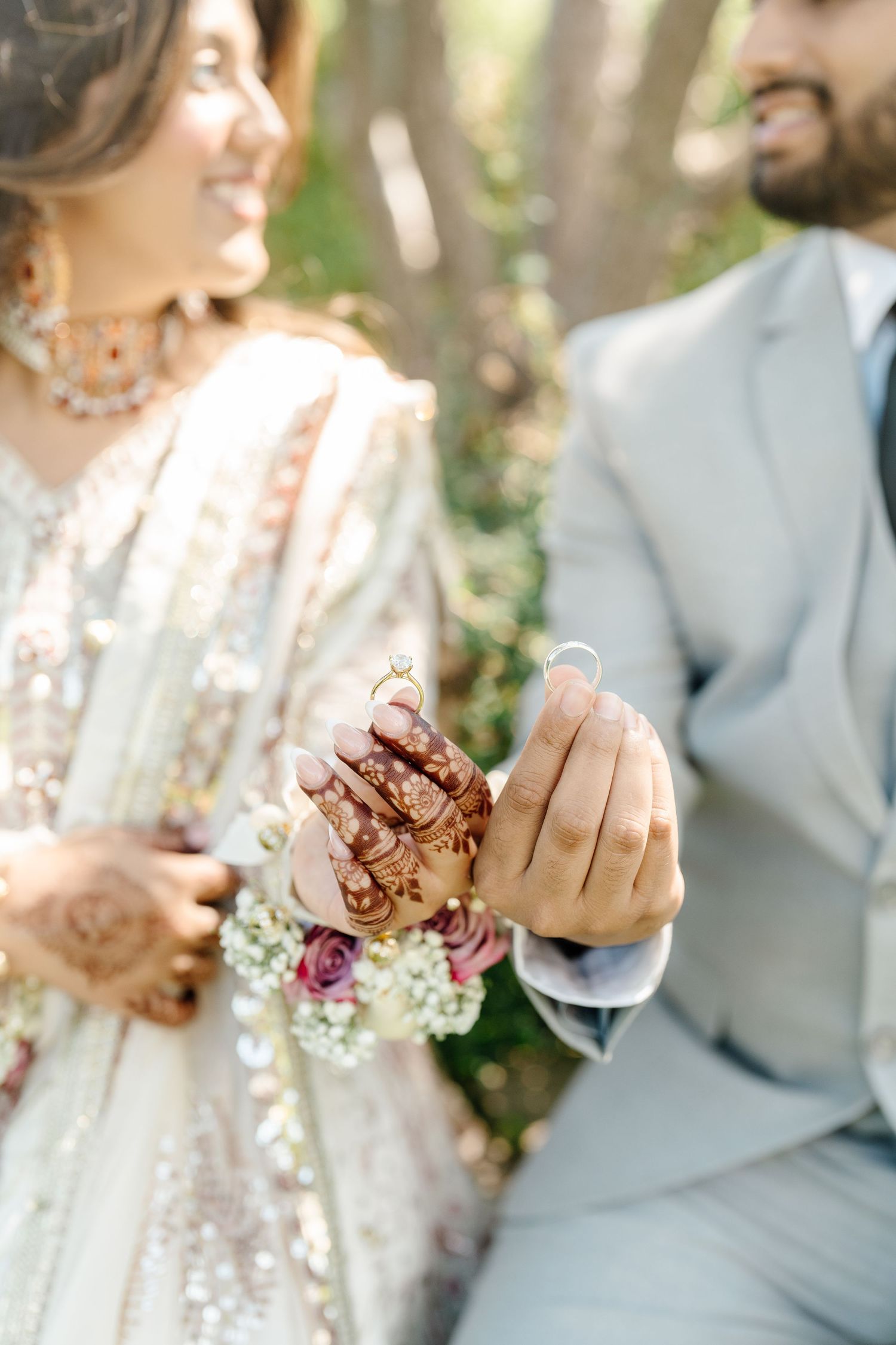 Indian bride and groom showing wedding rings, bride with henna hands holding floral bouquet outdoors.