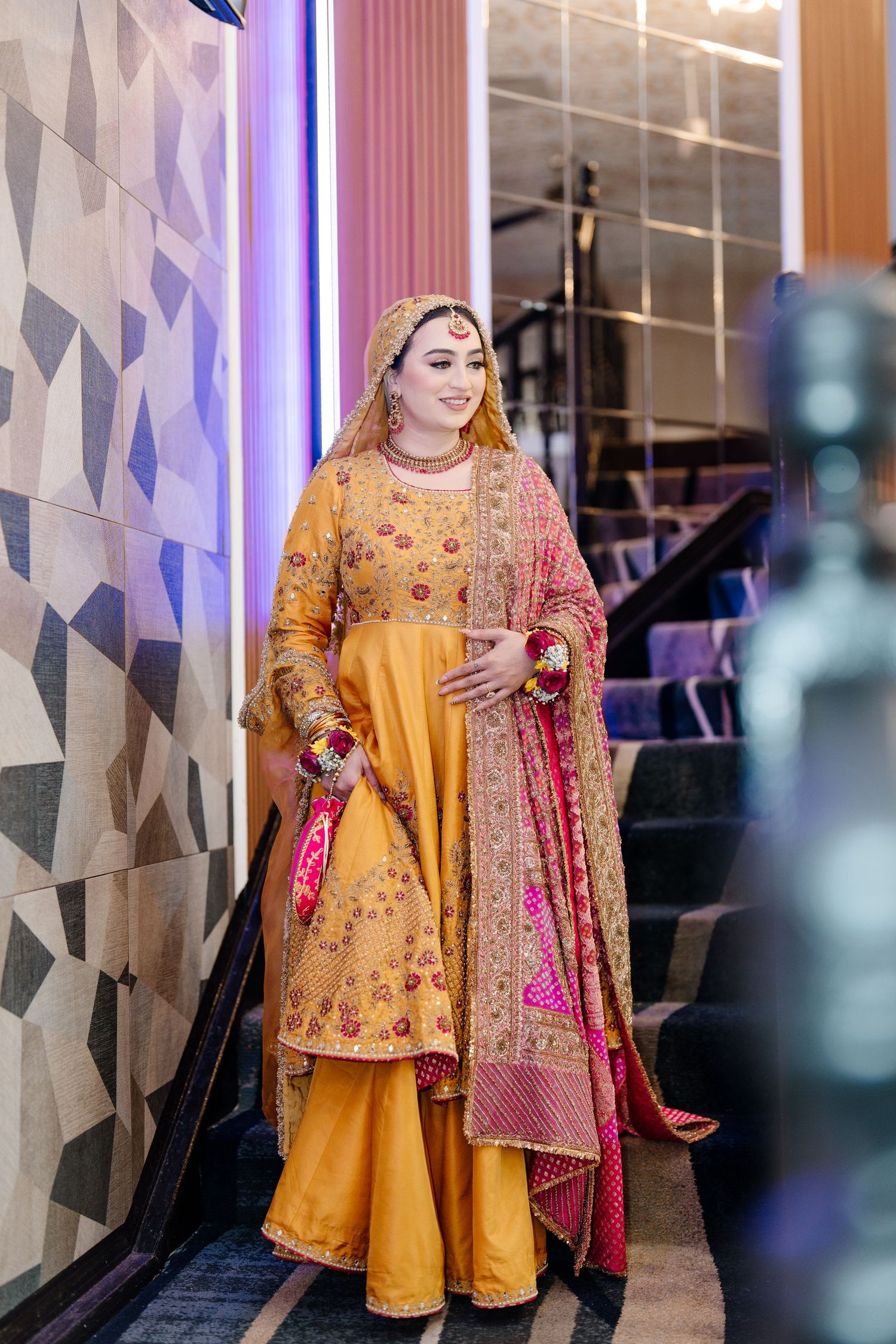South Asian bride in mustard yellow embroidered gharara with pink dupatta posing on decorative staircase.