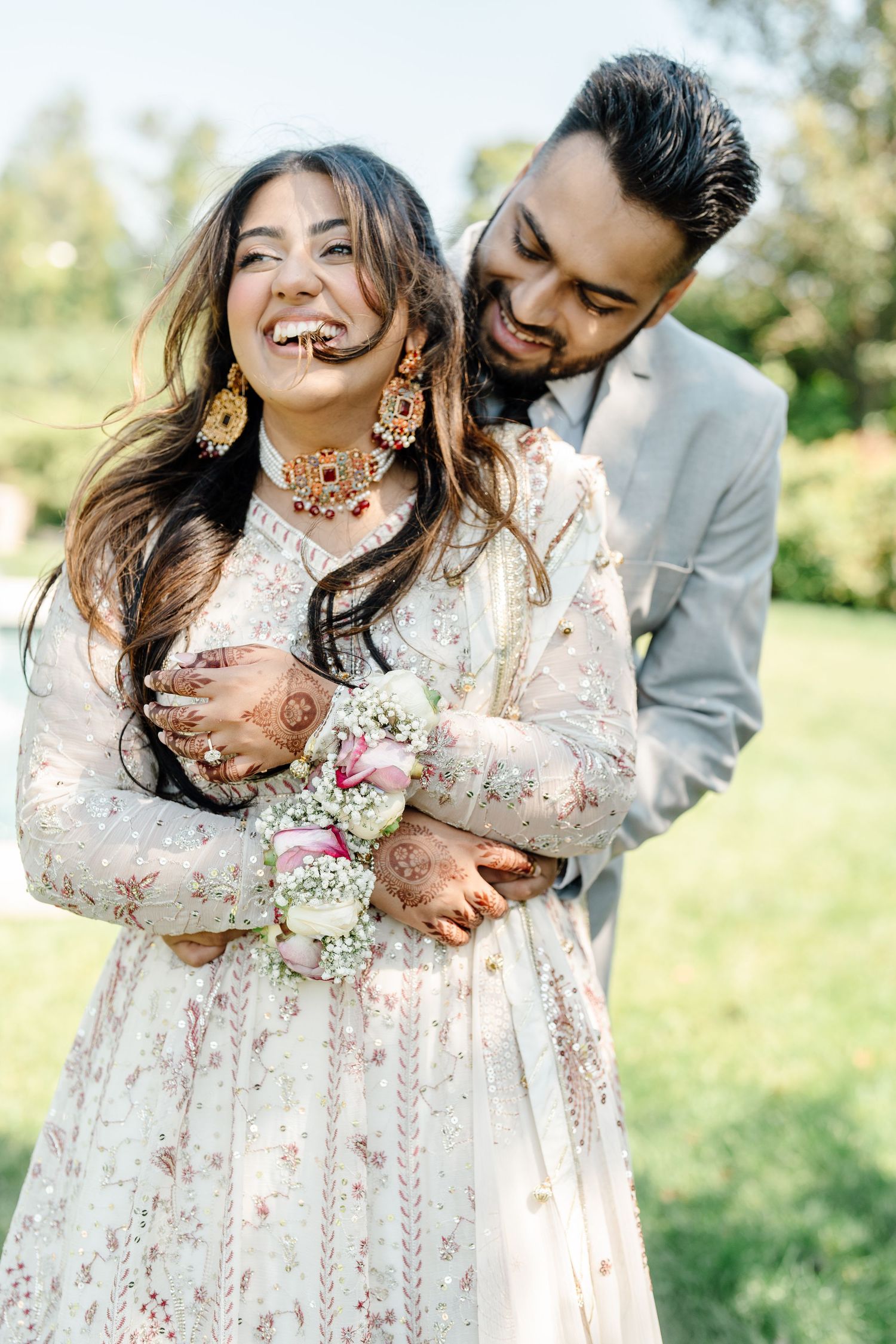 Joyful Indian couple embracing outdoors, bride in white embroidered lehenga with mehndi and gold earrings.