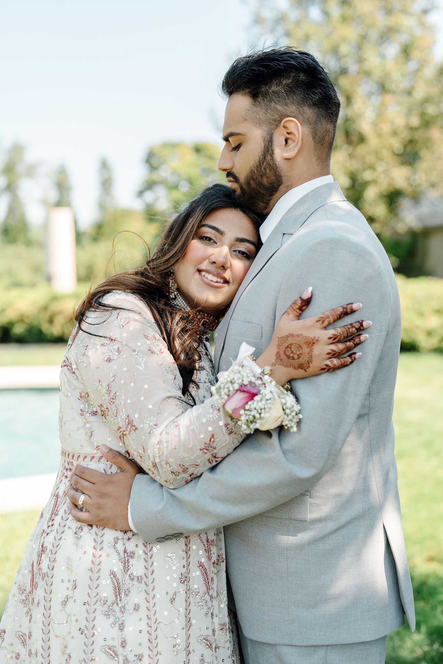 Indian bride and groom sharing tender embrace outdoors, bride smiling in embroidered lehenga with henna hands.