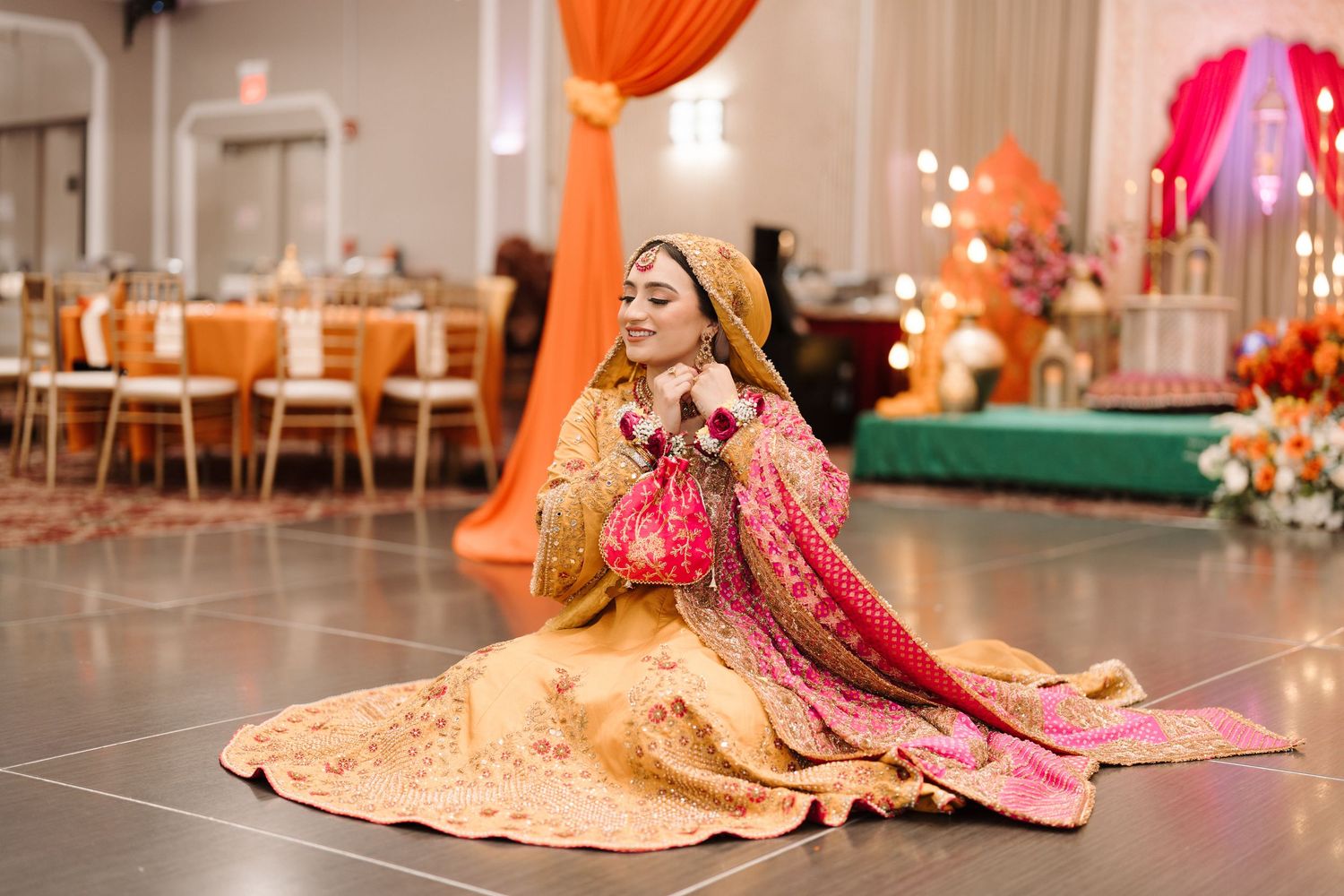 Pakistani bride in embroidered golden lehenga seated elegantly on dance floor with colorful wedding decor behind her.