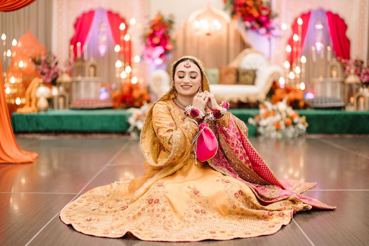 Bride in golden lehenga and pink dupatta sitting gracefully on dance floor at vibrant South Asian wedding ceremony.