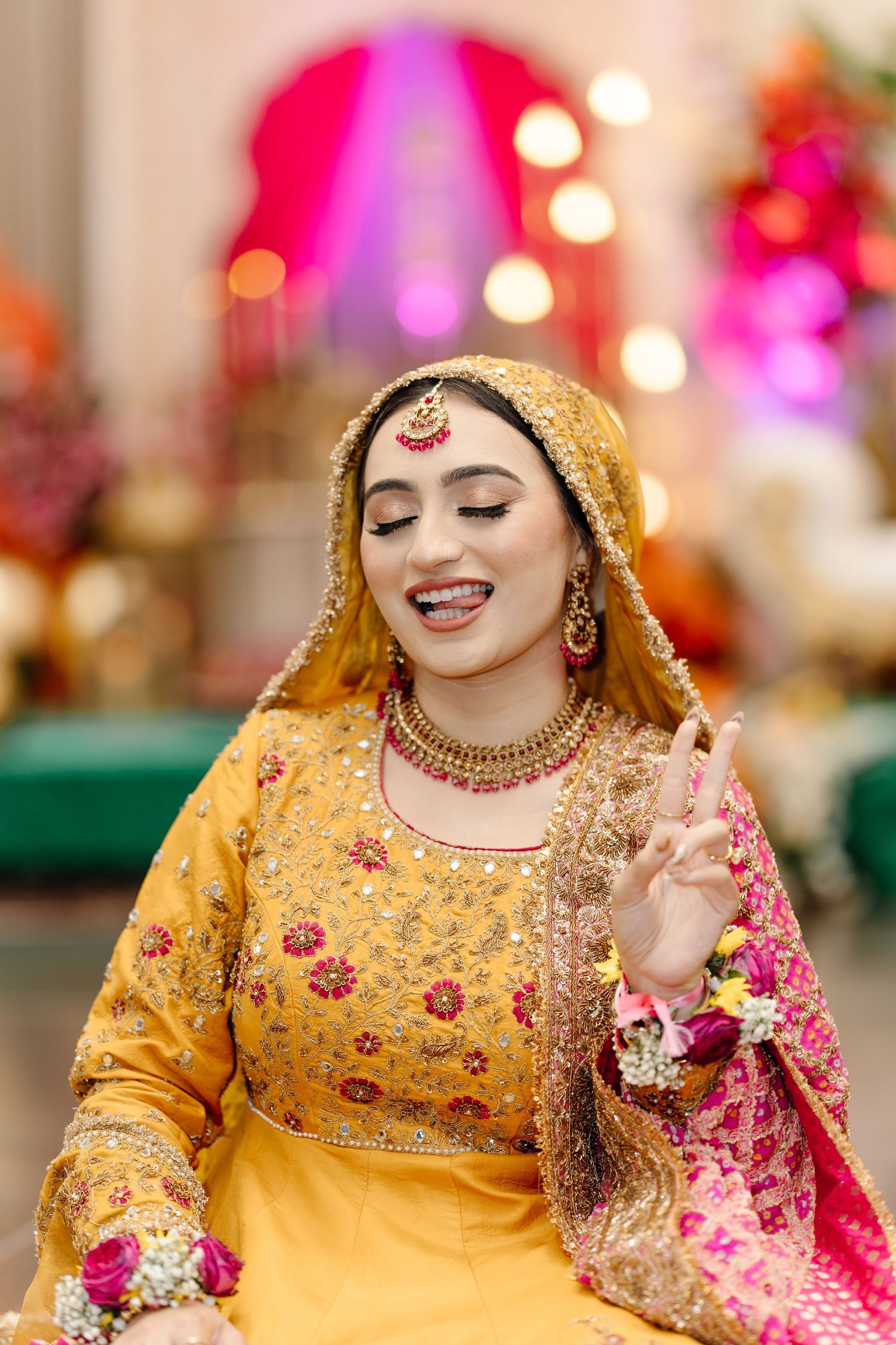 Smiling South Asian bride in yellow embroidered outfit with gold jewelry and floral accessories at mehndi ceremony.