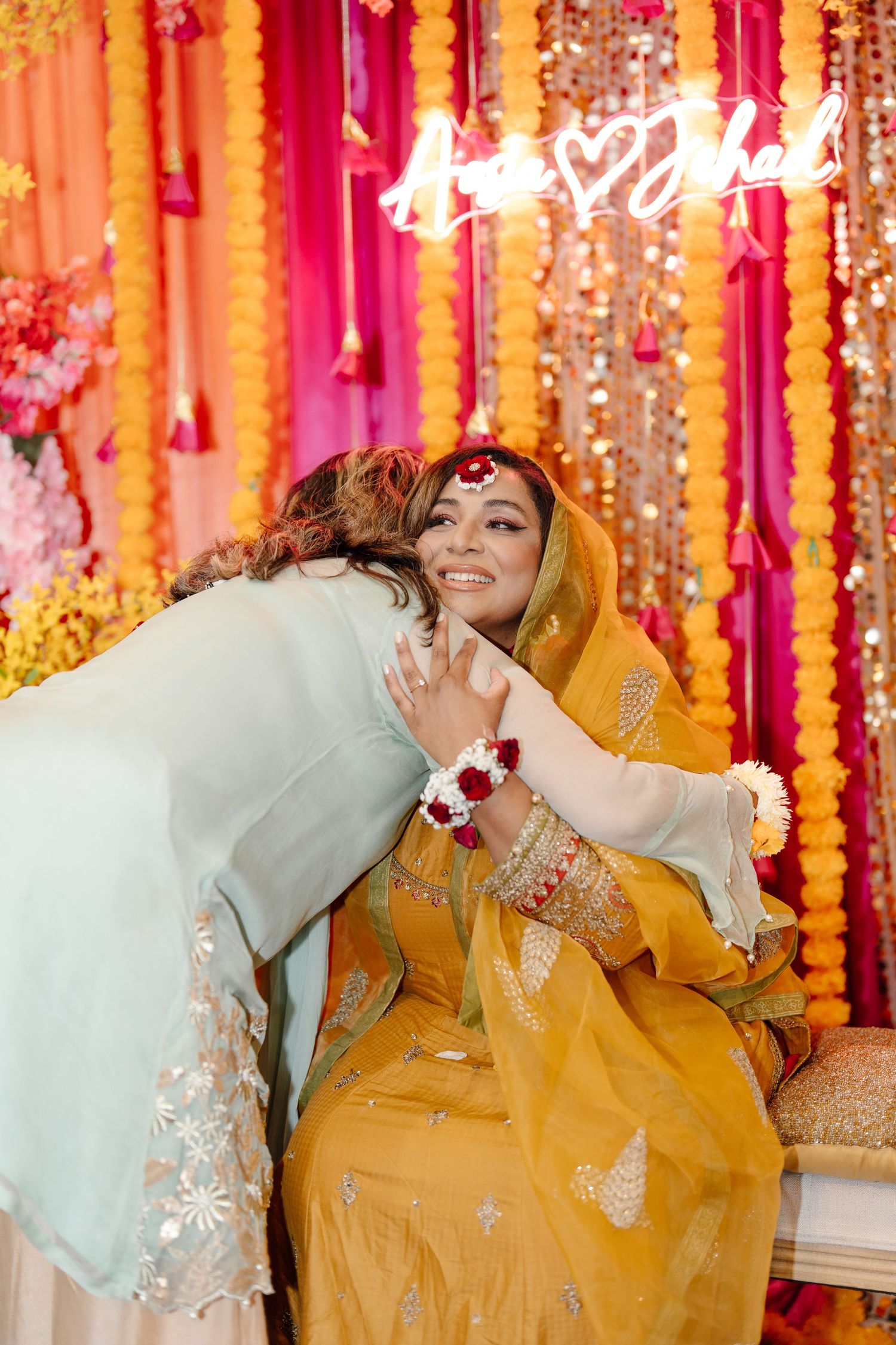 Bride in yellow lehenga smiling during emotional haldi ceremony embrace under colorful floral decorations.