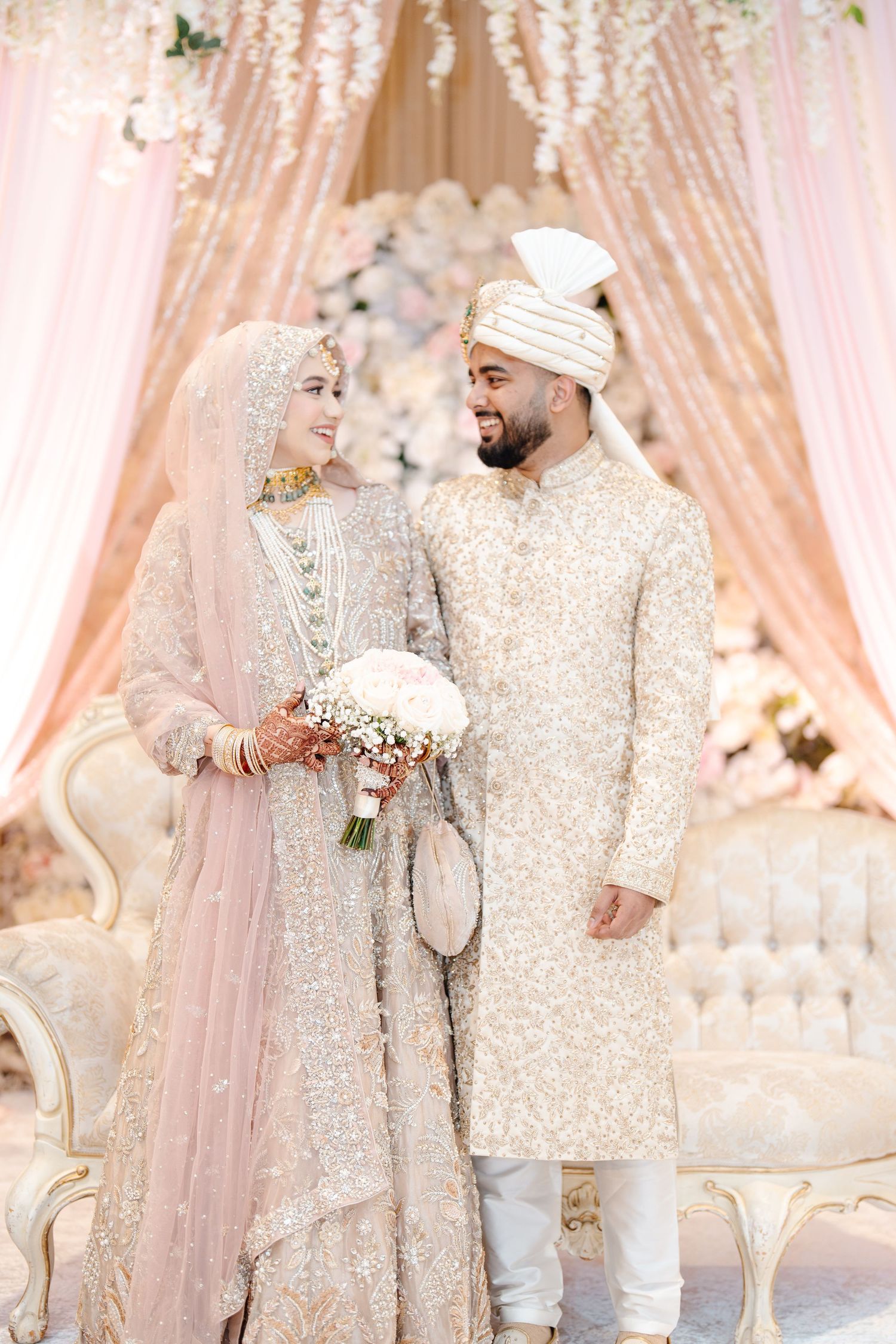 Muslim couple in matching ivory sequined wedding attire posing with bridal bouquet at reception.