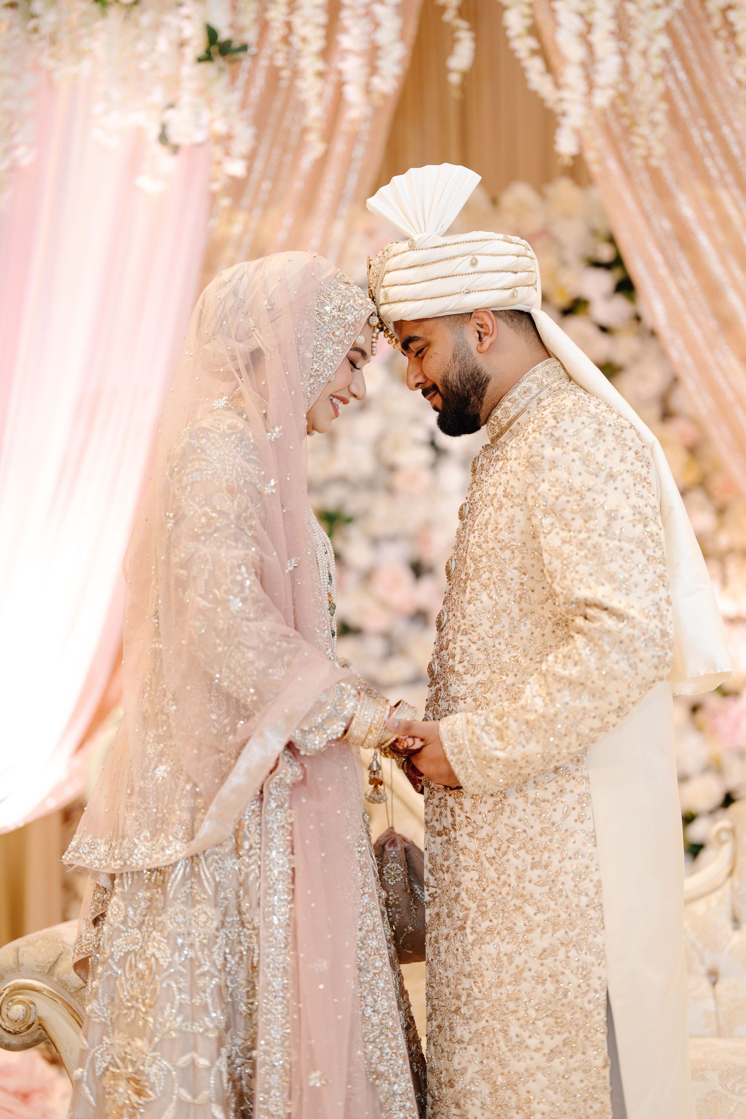 Muslim bride and groom in ivory and gold embroidered wedding attire standing under floral canopy.