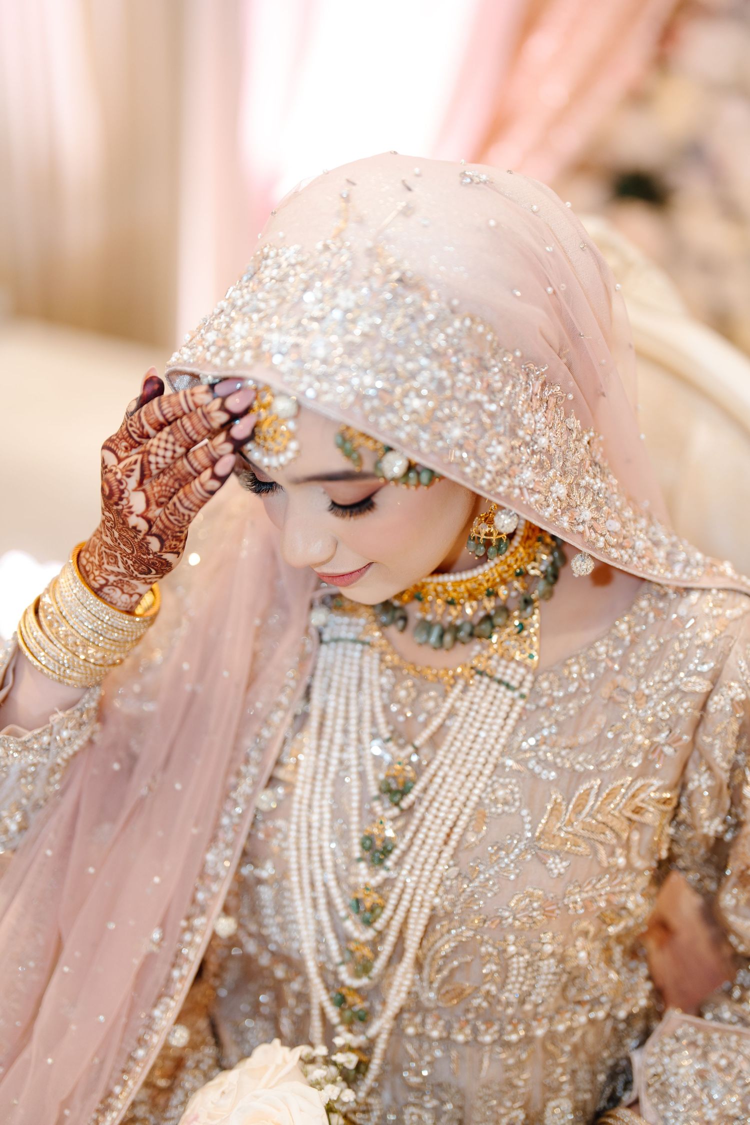 Pakistani Muslim bride in heavily embroidered ivory outfit with pearl jewelry and mehndi hands.