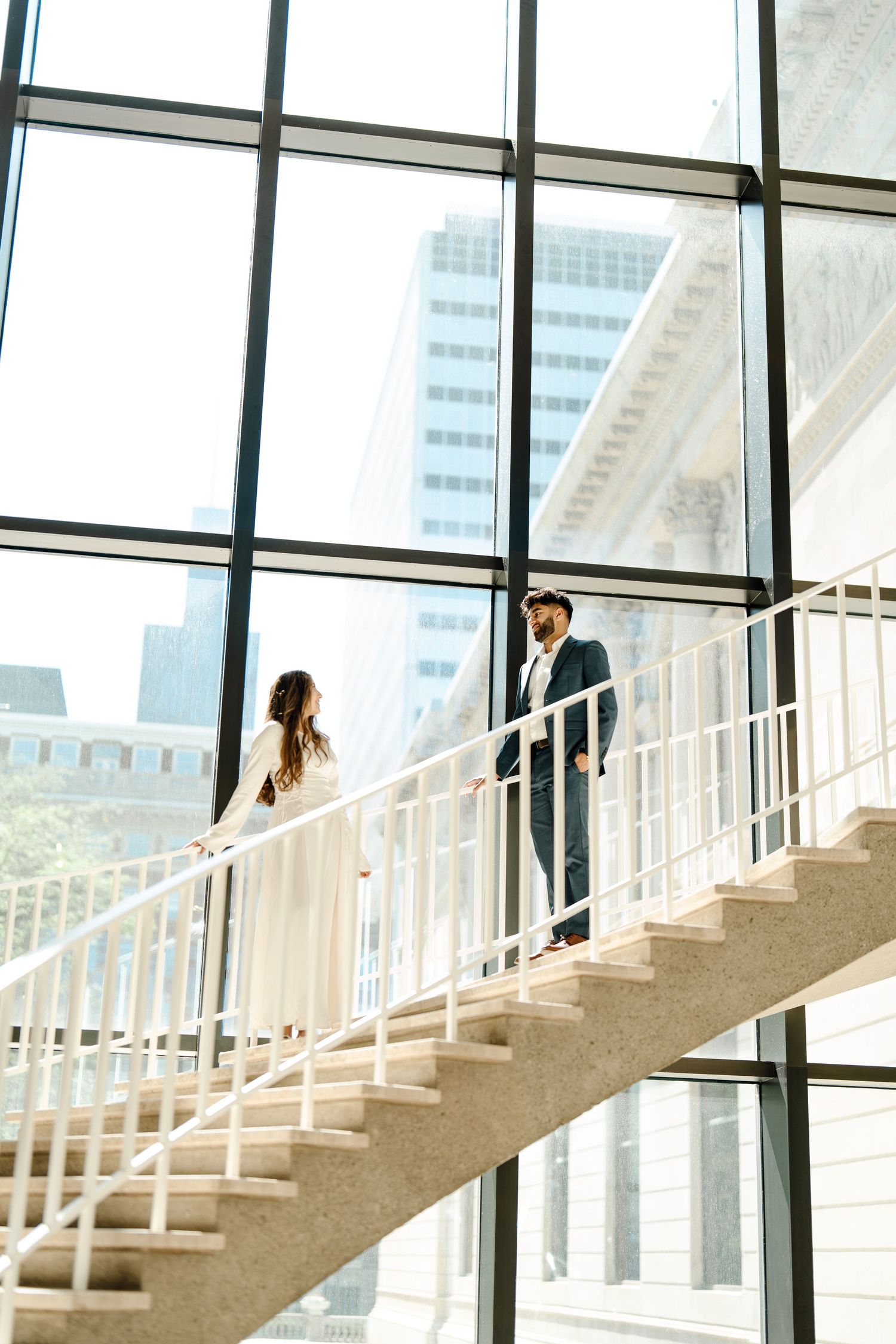 Bride and groom standing on modern staircase with floor-to-ceiling windows and city view.