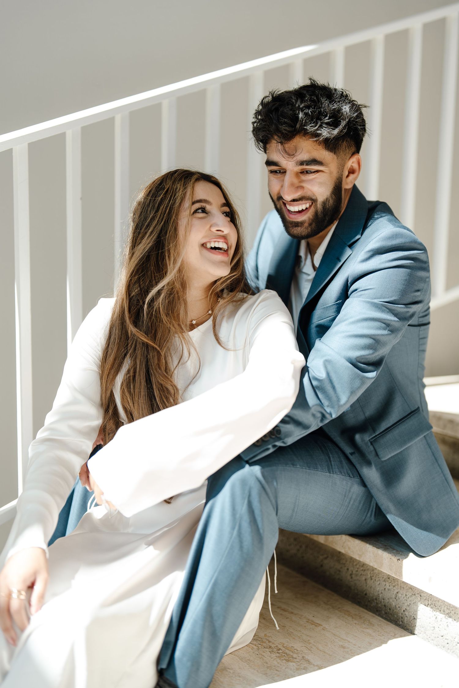 Happy couple laughing together while sitting on staircase steps in wedding attire.