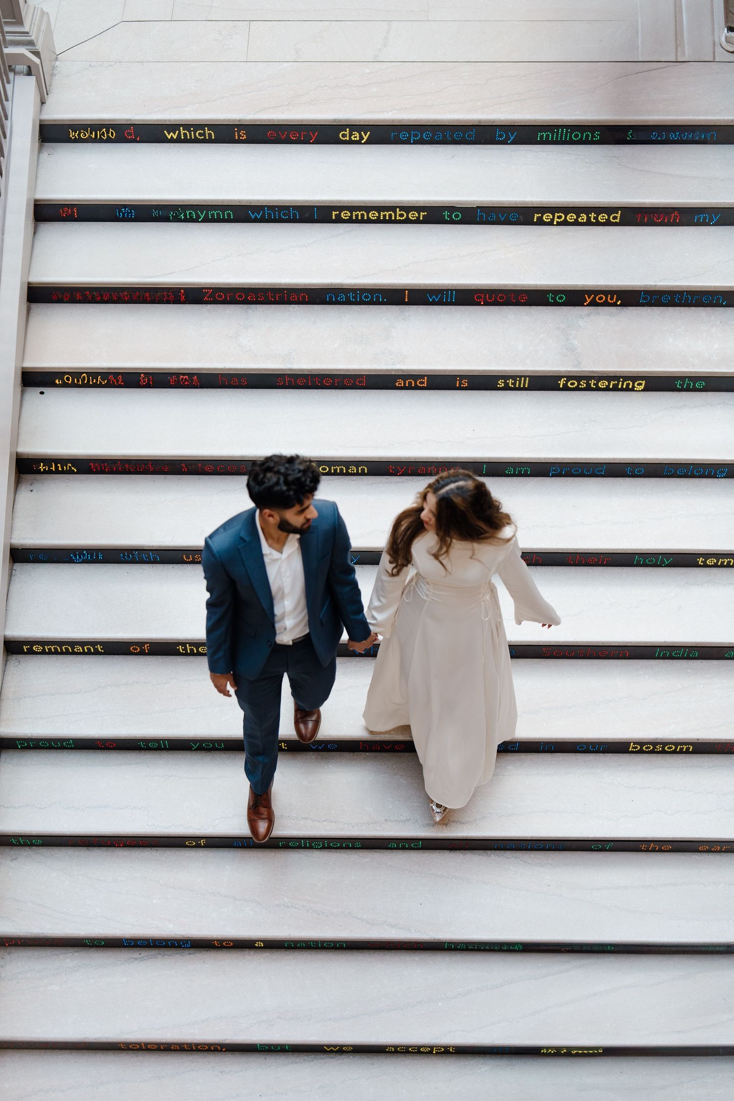 Aerial view of bride and groom holding hands walking down colorful text-inscribed stairs.