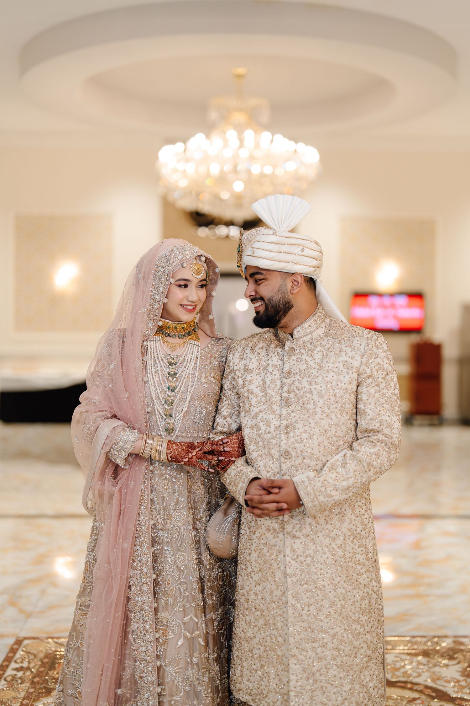 Pakistani bride and groom in ivory and blush embroidered wedding outfits smile lovingly at each other indoors.