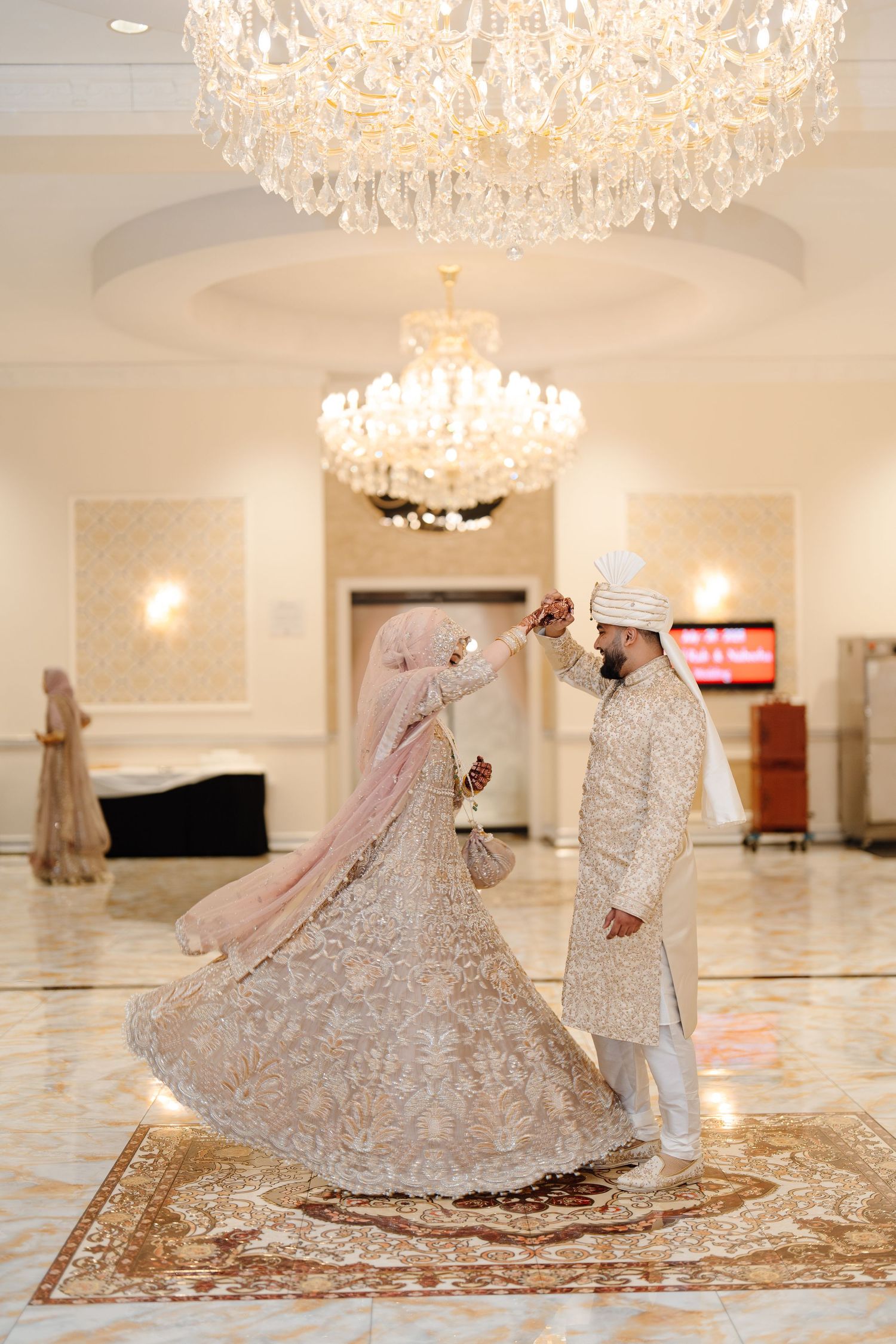 South Asian bride and groom share a romantic dance under a crystal chandelier in elegant traditional wedding attire.