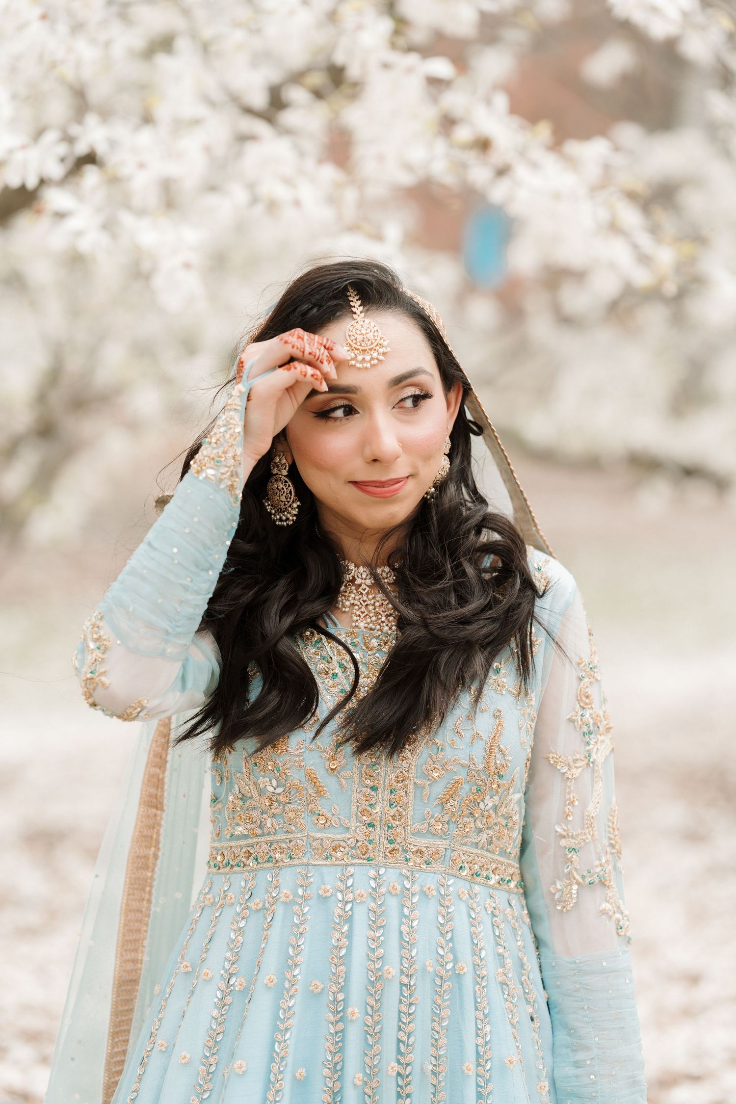 Indian bride in powder blue anarkali adjusting maang tikka jewelry among white cherry blossom trees.