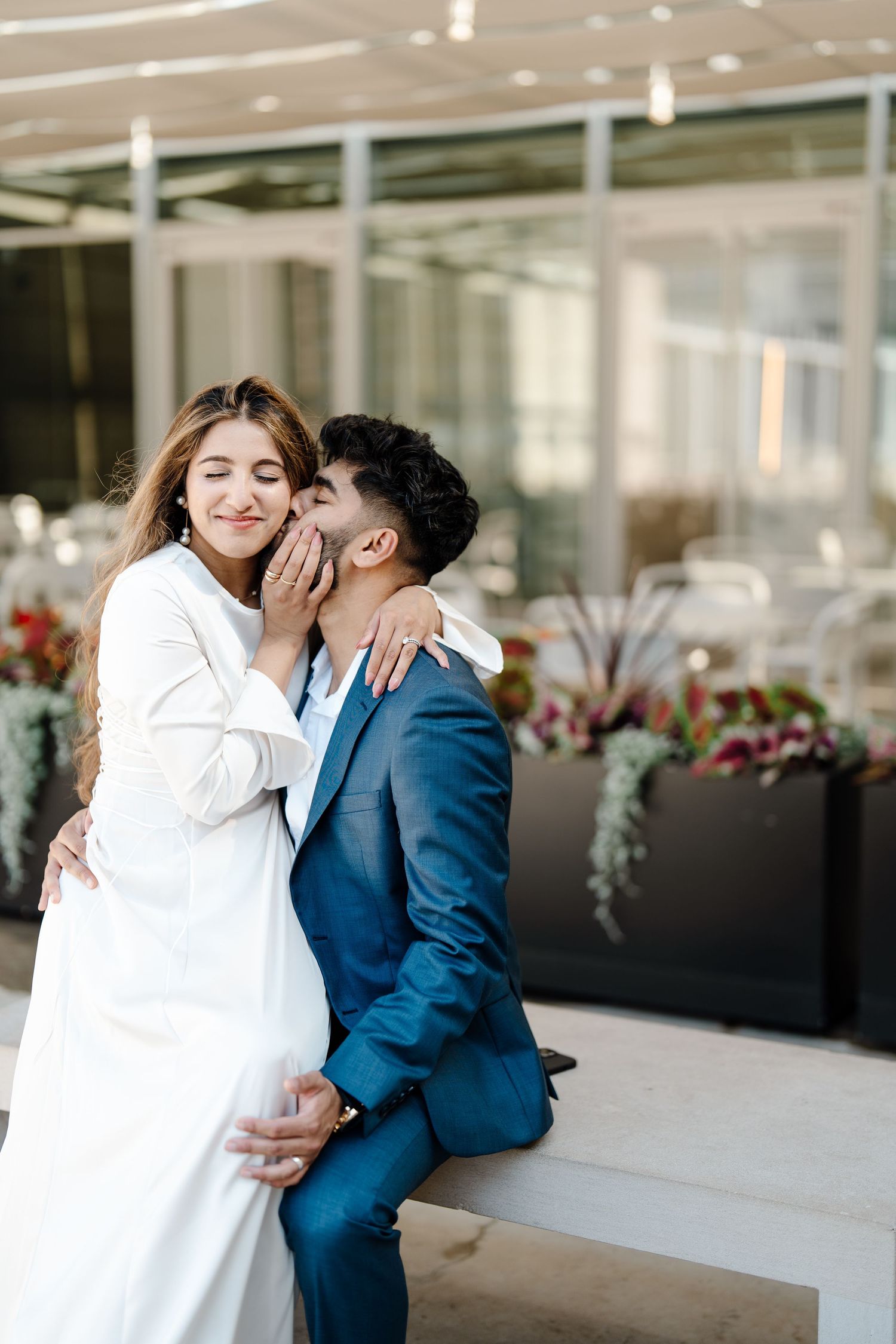 Groom in blue suit kissing bride on cheek while sitting outdoors at urban venue.