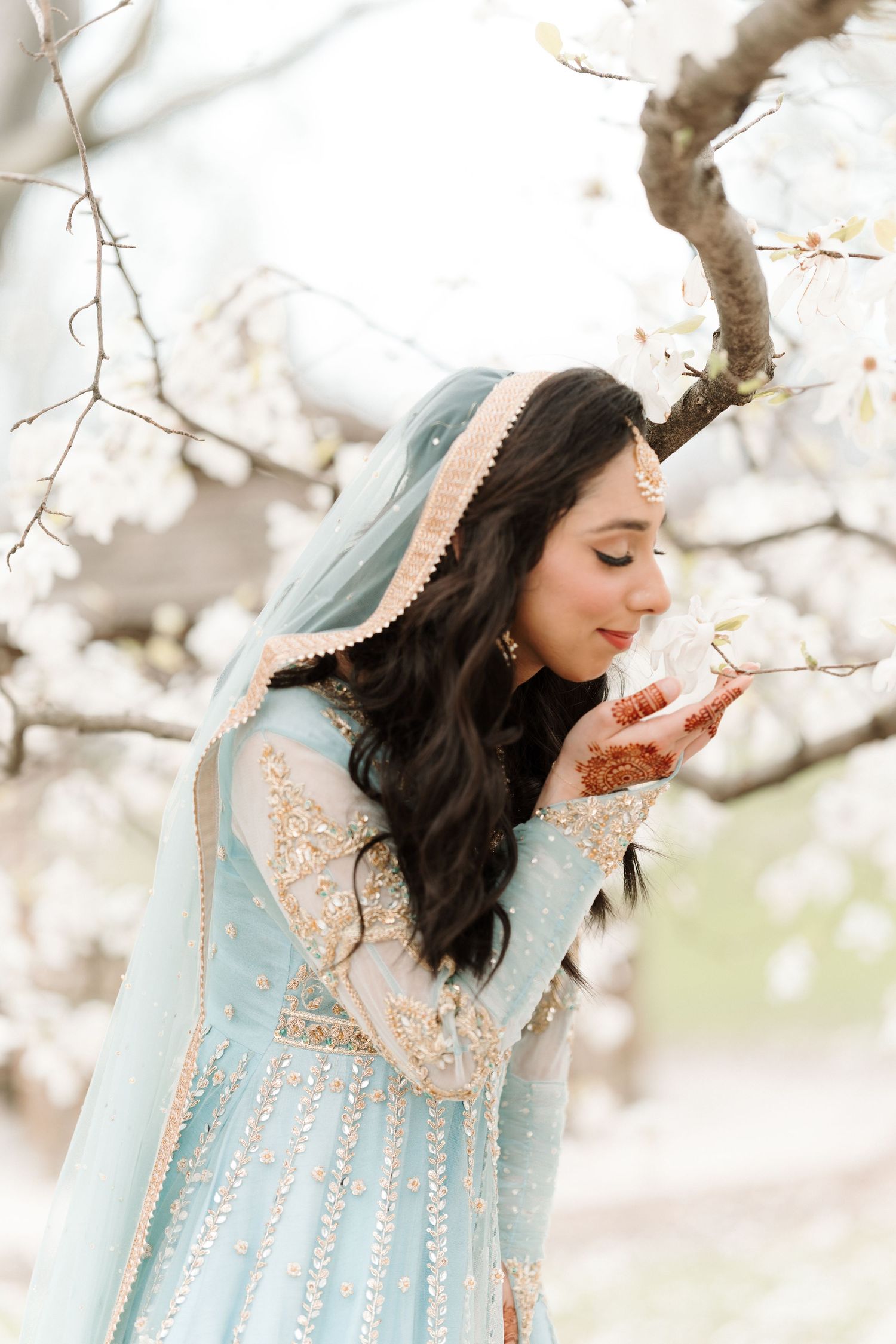 Indian bride in light blue bridal outfit smelling white cherry blossoms with mehndi-adorned hands.