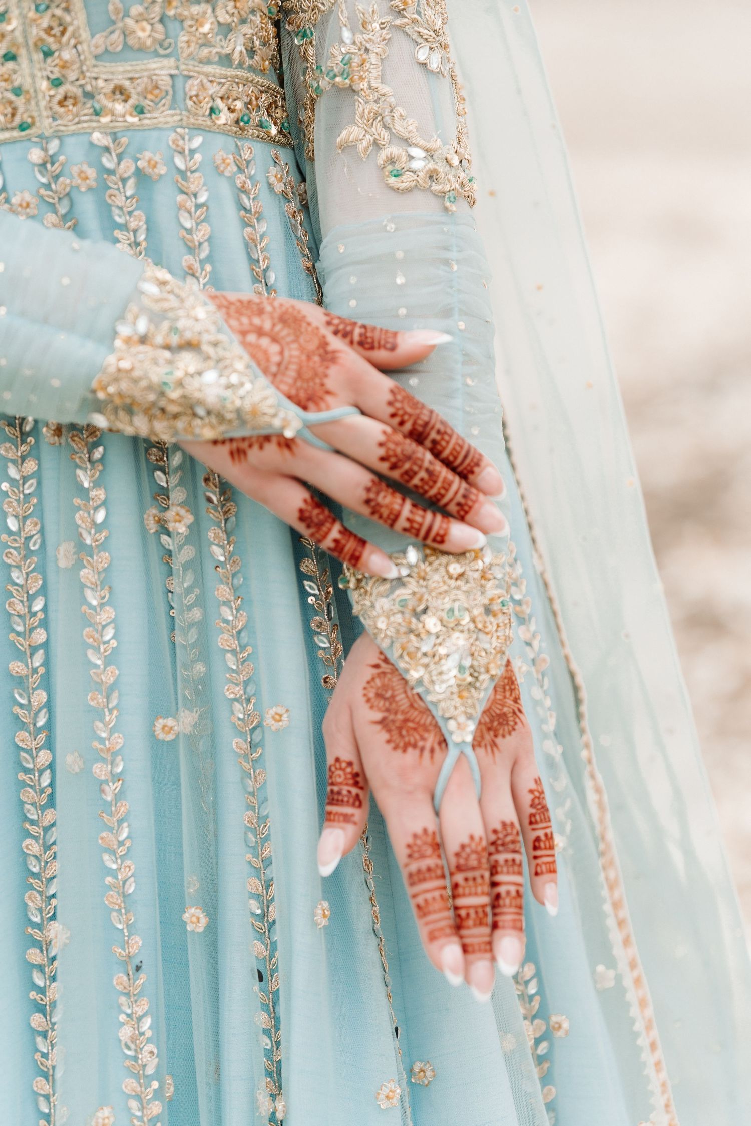 Close-up of bridal hands adorned with intricate henna mehndi designs against a light blue embroidered lehenga.