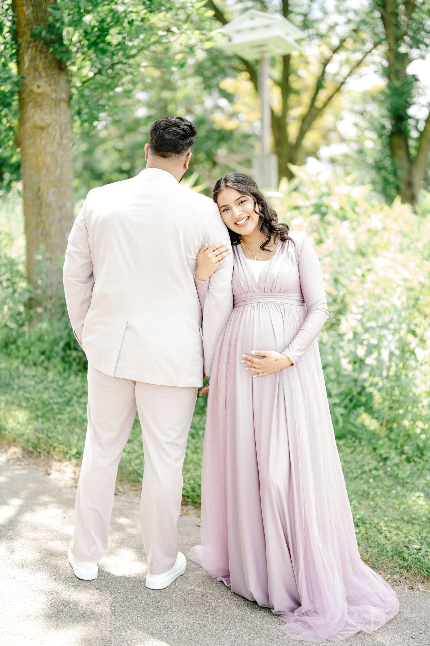 Pregnant woman in lavender gown smiling beside partner in cream suit in green park.