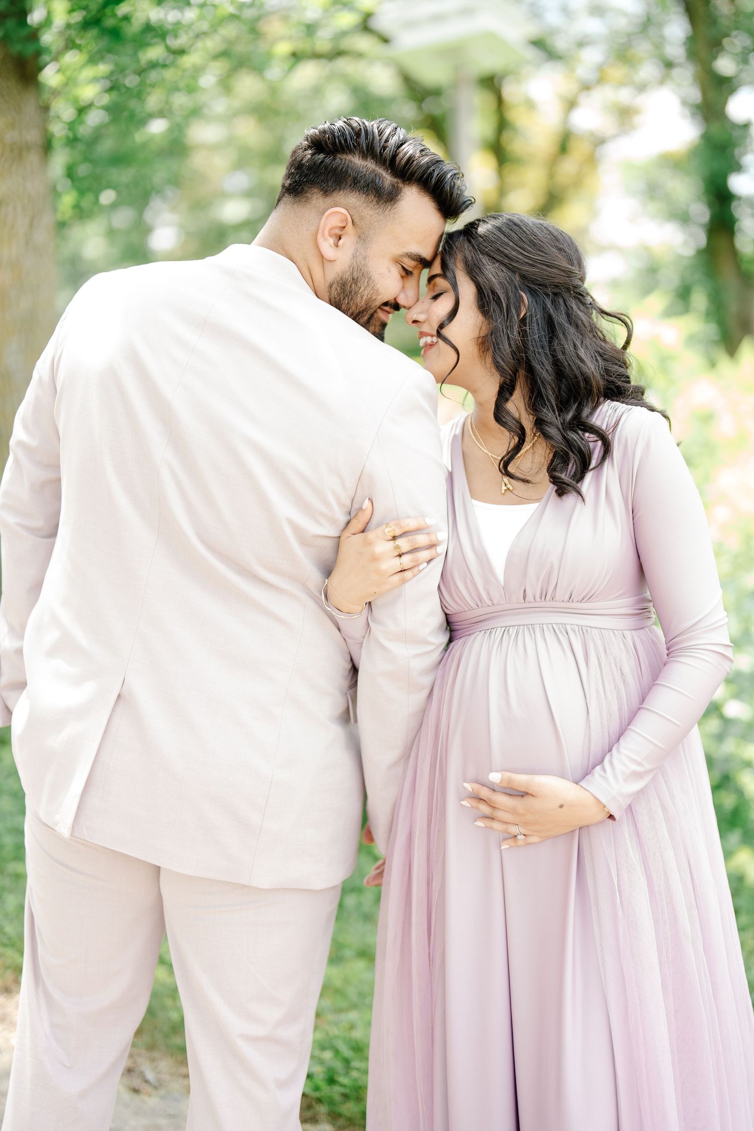 Expecting couple in soft pink and lavender outfits sharing tender forehead touch outdoors.
