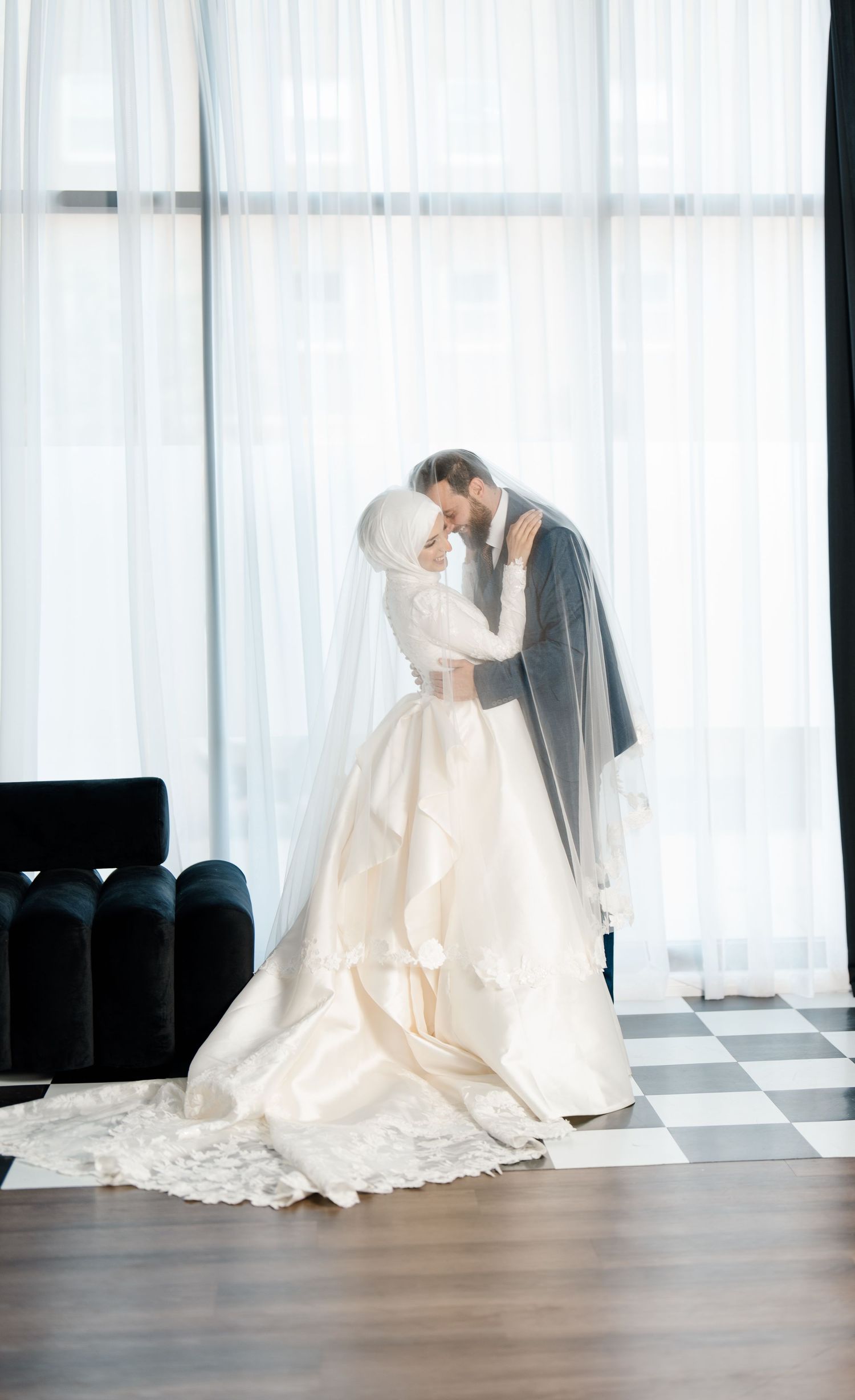 Muslim bride and groom sharing romantic moment under veil in white ball gown by bright window curtains.