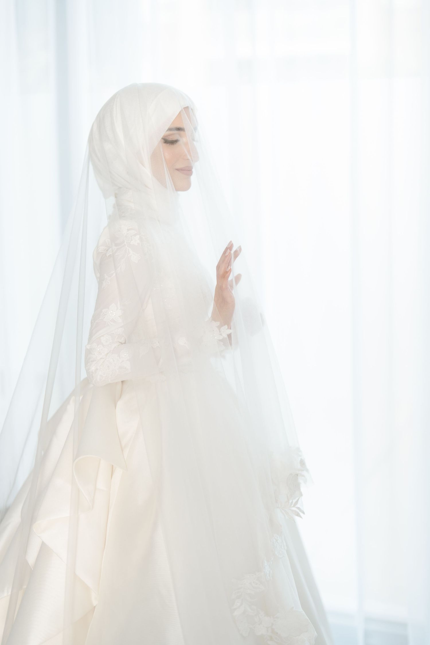 Muslim bride in white hijab and lace ball gown standing gracefully by sheer white curtained window.