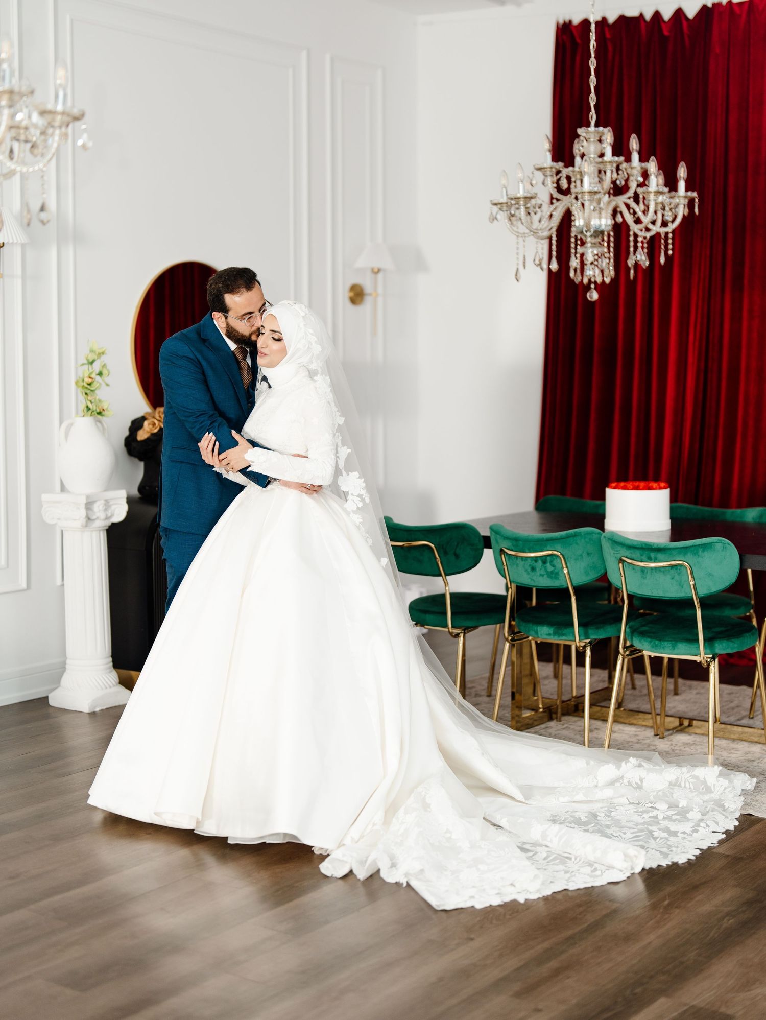 Muslim couple in elegant wedding attire posing in white room with red velvet curtains and green chairs.