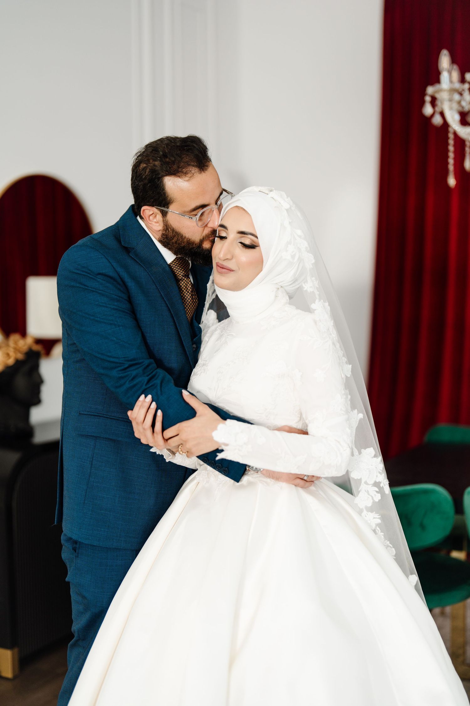 Muslim groom in navy suit kissing bride in white hijab wedding dress in elegant red and white venue.