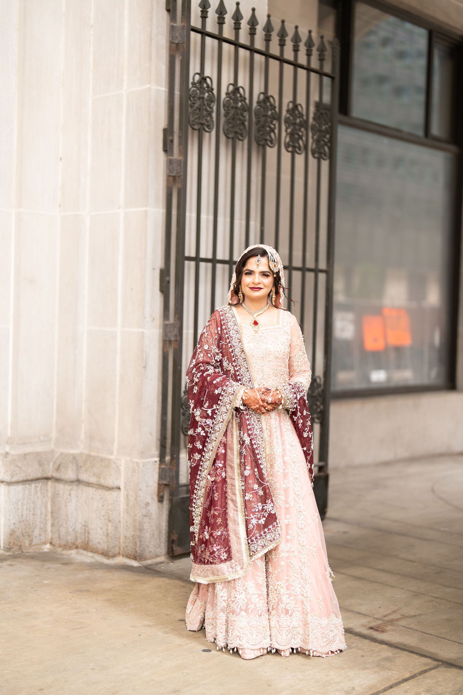 South Asian bride poses in pink embroidered lehenga with burgundy dupatta outside a stone building.