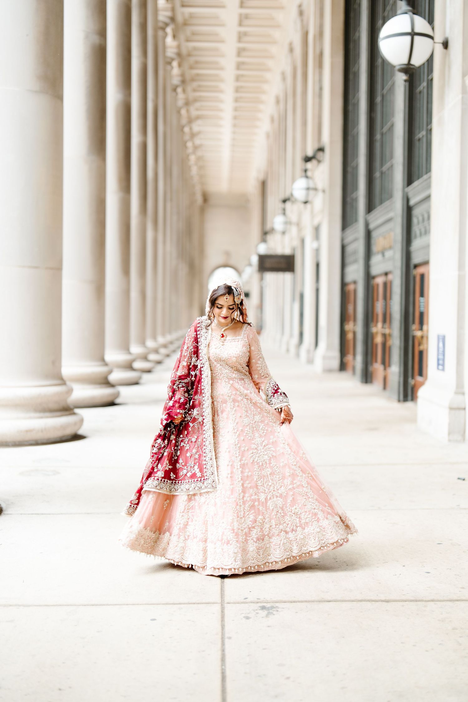 South Asian bride twirls in blush pink embroidered lehenga with red dupatta between grand stone columns.