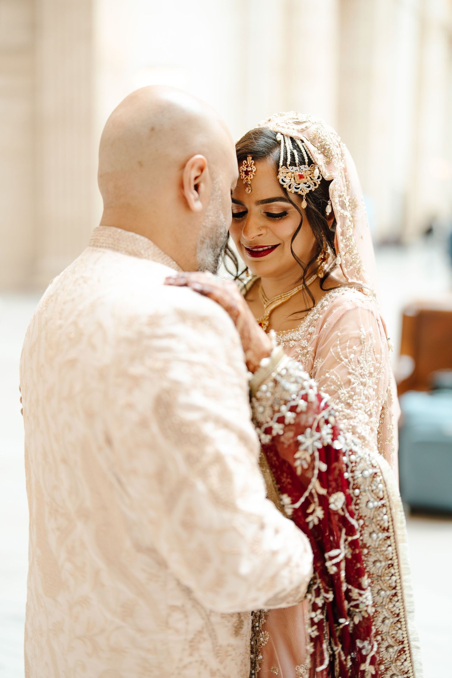 South Asian groom in cream sherwani tenderly holds bride in red and gold lehenga with ornate bridal jewelry.